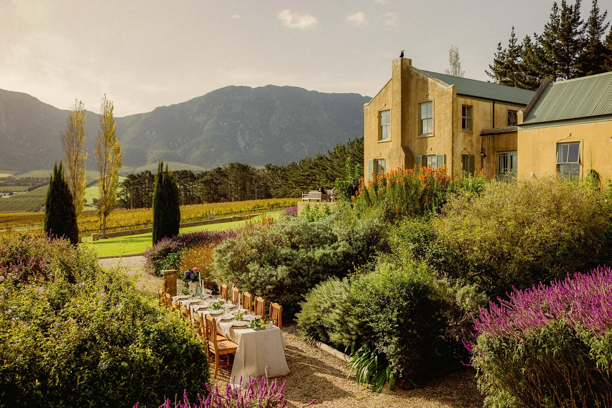 A garden setting with a long dining table decorated for an outdoor meal, surrounded by colorful flowers and lush greenery. Private luxury villa (accommodation) in Hemel-en-Aarde, Hermanus.
