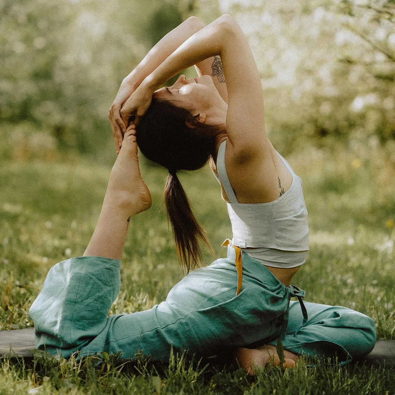 A woman practicing yoga outdoors on grass, performing a backbend pose with her hands holding her feet, wearing a white tank top and green pants. Private luxury villa (accommodation) in Hemel-en-Aarde, Hermanus.