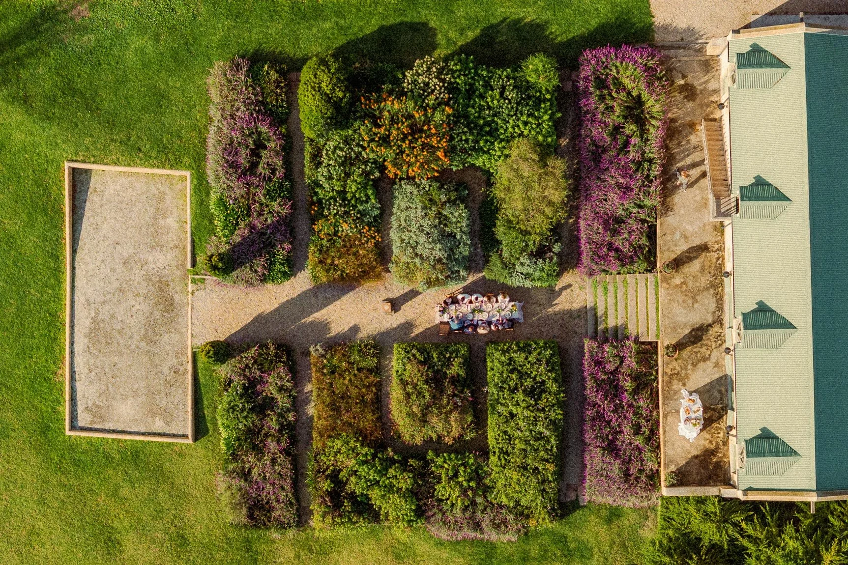 Aerial view of a garden with various colorful flowering bushes and trees, a gravel patio area with a table of people, and a building with a green roof on the right side. Private luxury villa (accommodation) in Hemel-en-Aarde, Hermanus.