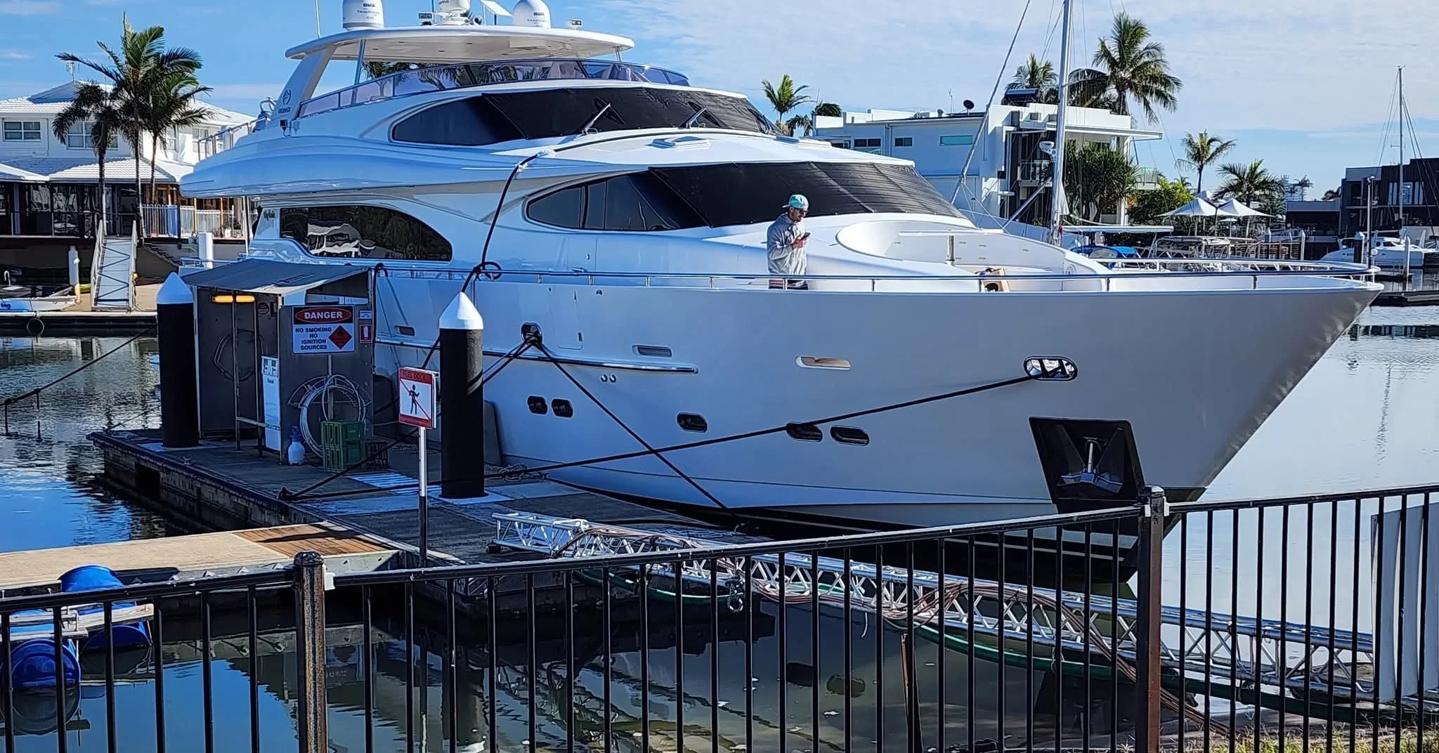 A large white luxury yacht docked at a marina with palm trees and modern buildings in the background. There is a person on the yacht, and the marina has various boats and a floating platform.