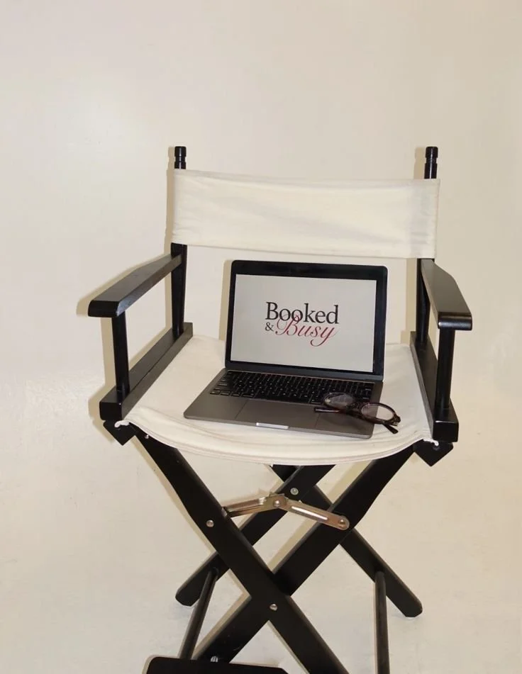 A director's chair with a laptop on the seat, displaying the text 'Booked & Busy', and a pair of eyeglasses beside the laptop.
