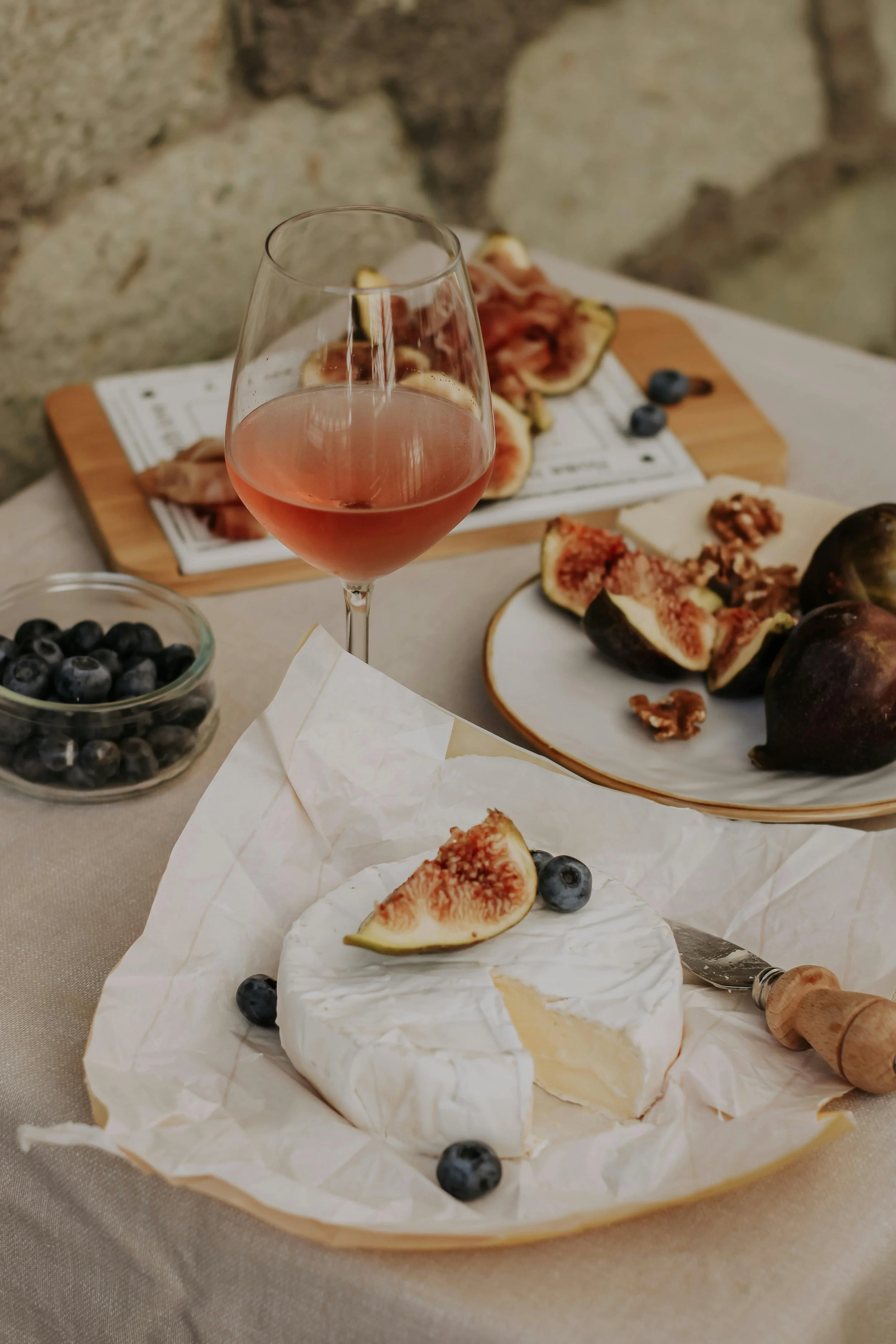 A table set with raw cheeses, fresh figs, blueberries, walnuts, a glass of pink wine, and a charcuterie board with cured meats and cheeses, against a rustic stone wall background.
