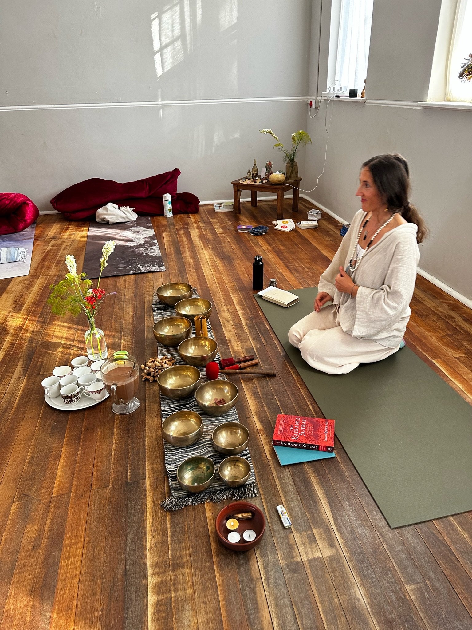 A woman sits on a yoga mat in a room decorated with spiritual objects, preparing for a sound bath session with brass singing bowls, tea cups, candles, and books on meditation and spirituality.