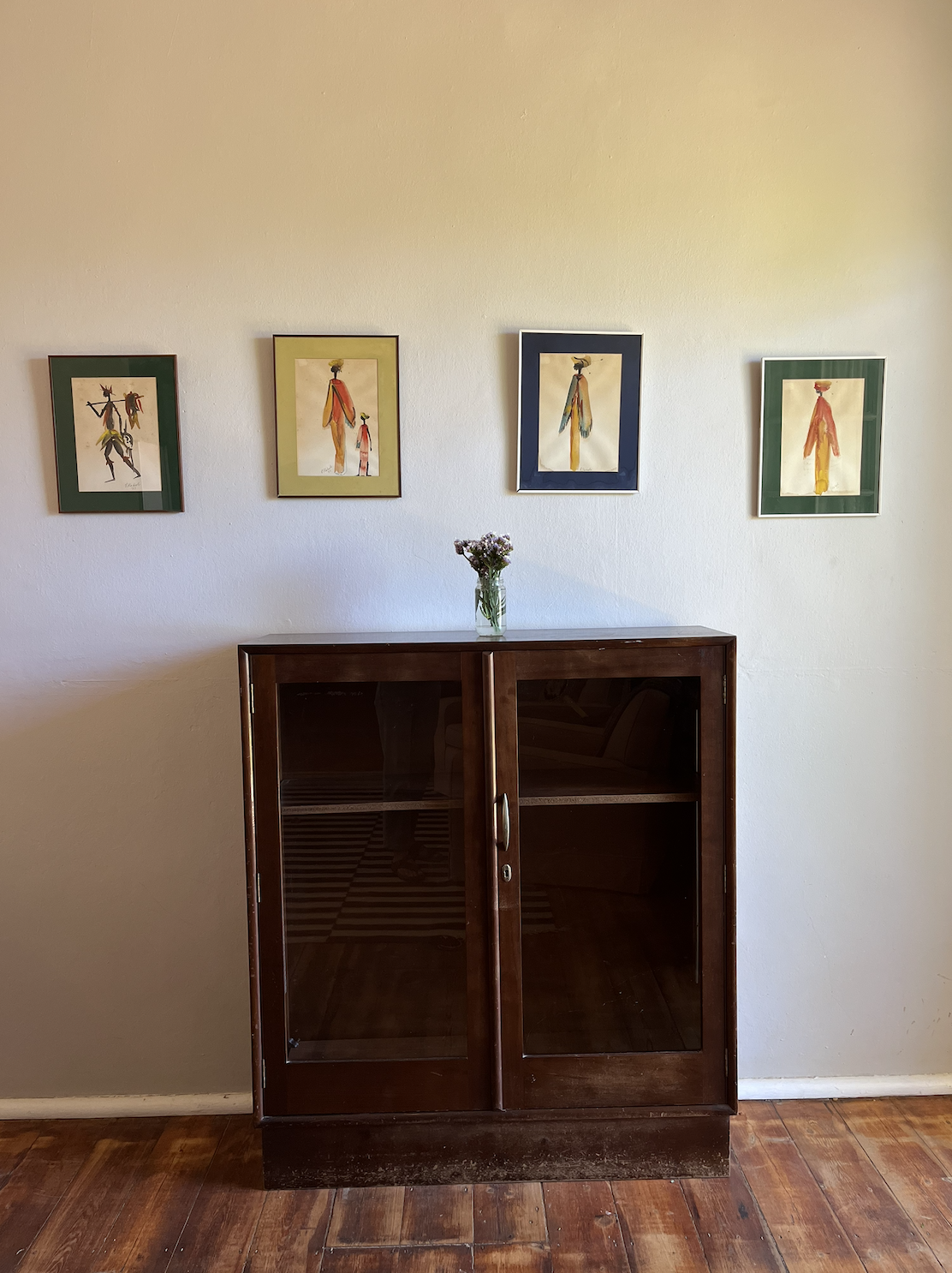 A wooden cabinet with glass doors is placed against a wall. On top of the cabinet, there is a vase with flowers. Above the cabinet, four framed colorful abstract art prints are hung on the wall.