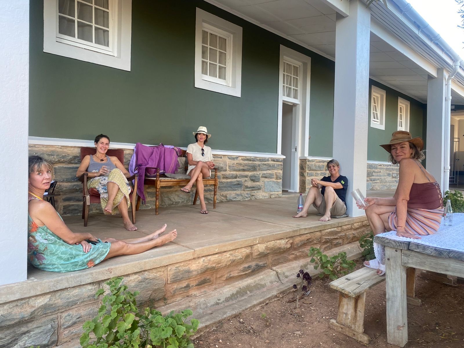 Five women are sitting on the porch of a house, some on chairs and others on the ground, enjoying a relaxed outdoor gathering. They are dressed casually, some with hats, and there are water bottles nearby.