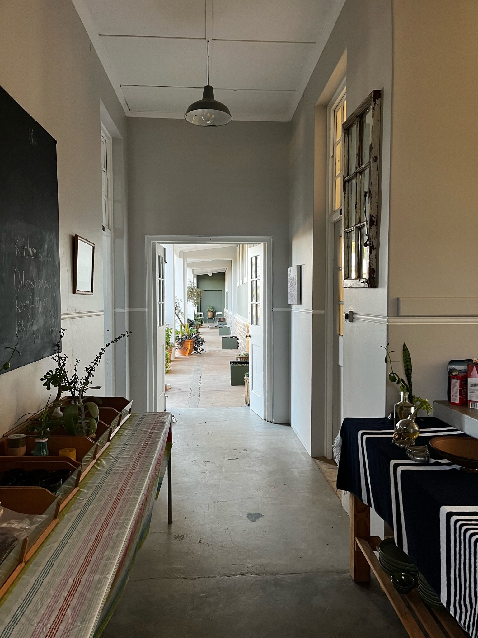 Inside view of a hallway with tables displaying plants and items, leading to a bright outdoor area with potted plants and furniture.
