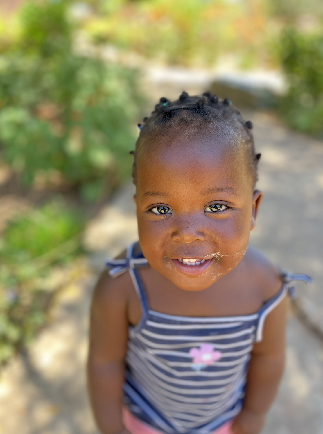 A young girl with braided hair and a striped tank top smiling at the camera outdoors with greenery in the background.
