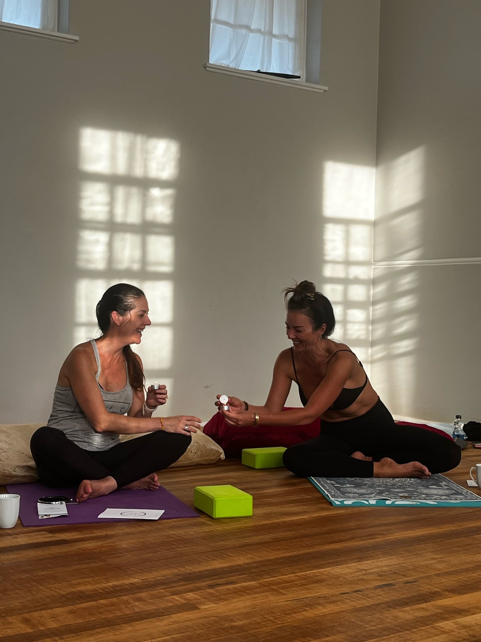 Two women sitting on yoga mats indoors, smiling and laughing, engaging in a conversation or activity, with sunlight casting shadows on the wall behind them.