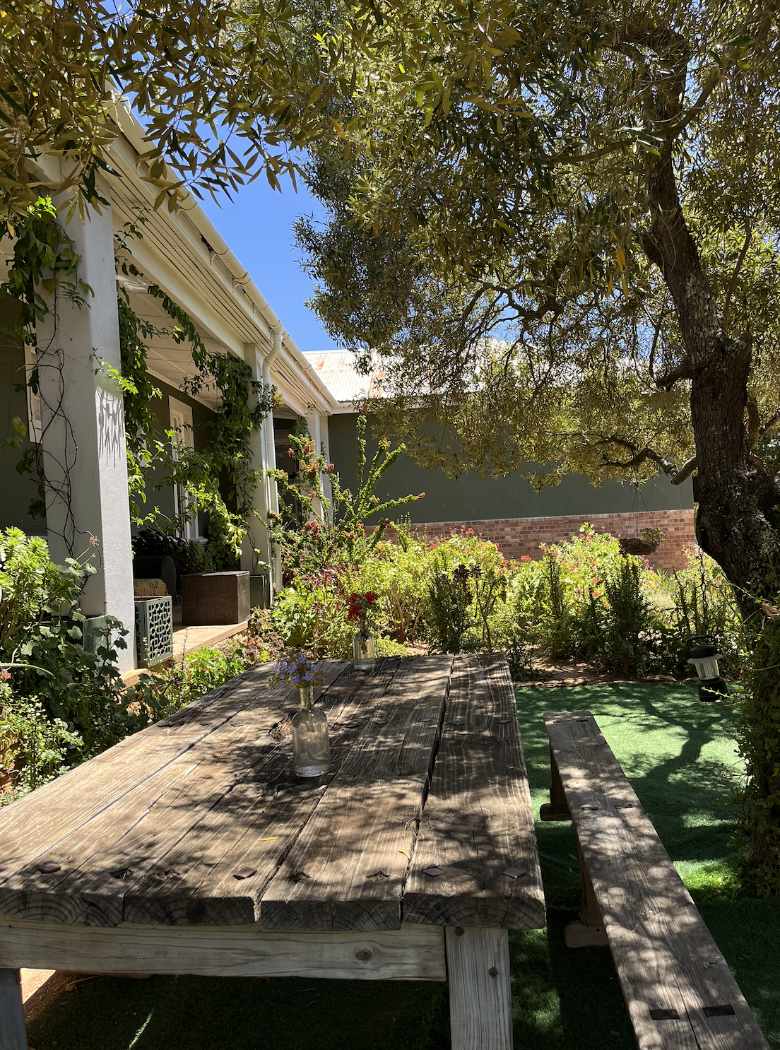 A rustic outdoor wooden table with vases holding flowers, surrounded by lush greenery and trees, under a shady garden setting beside a house with a porch.