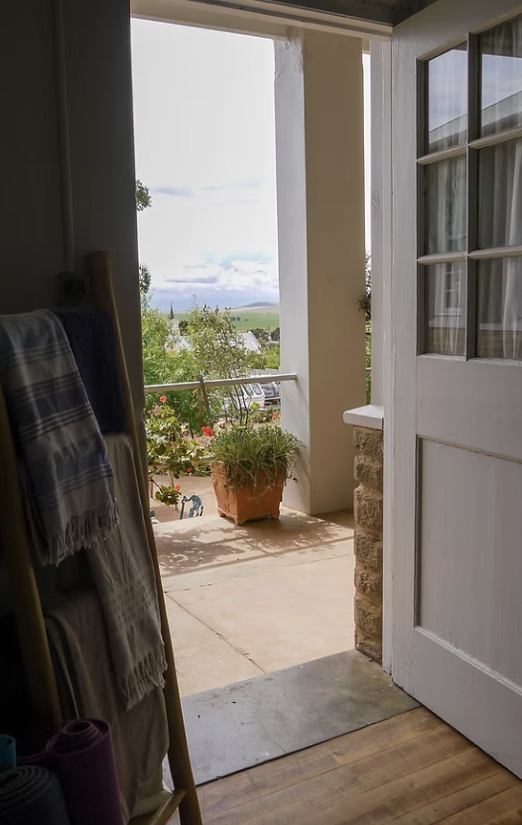 Open door leading to a balcony with a flower pot and scenic view of countryside with trees, fields, and cloudy sky.
