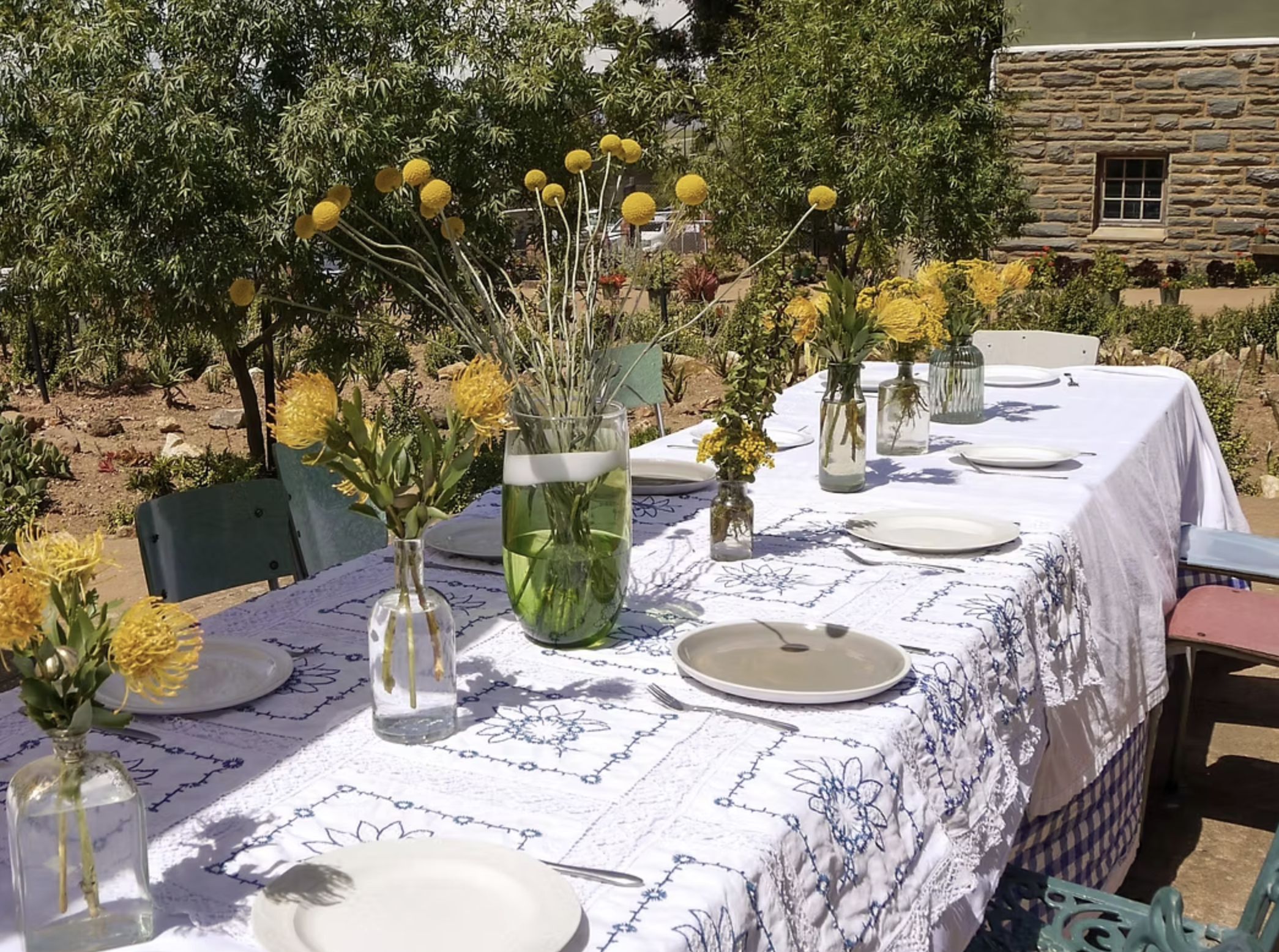 A long outdoor table set with white plates, cutlery, and decorated with multiple flower arrangements in clear and green glass vases on a white tablecloth with blue embroidery, under a sunny sky in a garden.