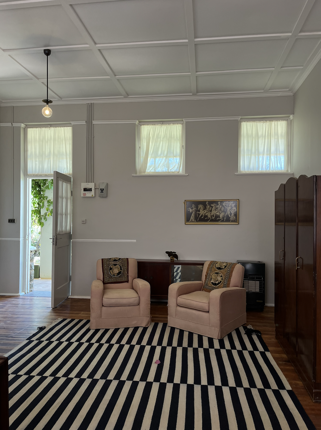 A living room with two pink armchairs, a black-and-white striped rug, wooden flooring, and a wooden cabinet. There's a door open to the outside, three small windows with curtains, a framed artwork on the wall, and a hanging lightbulb.