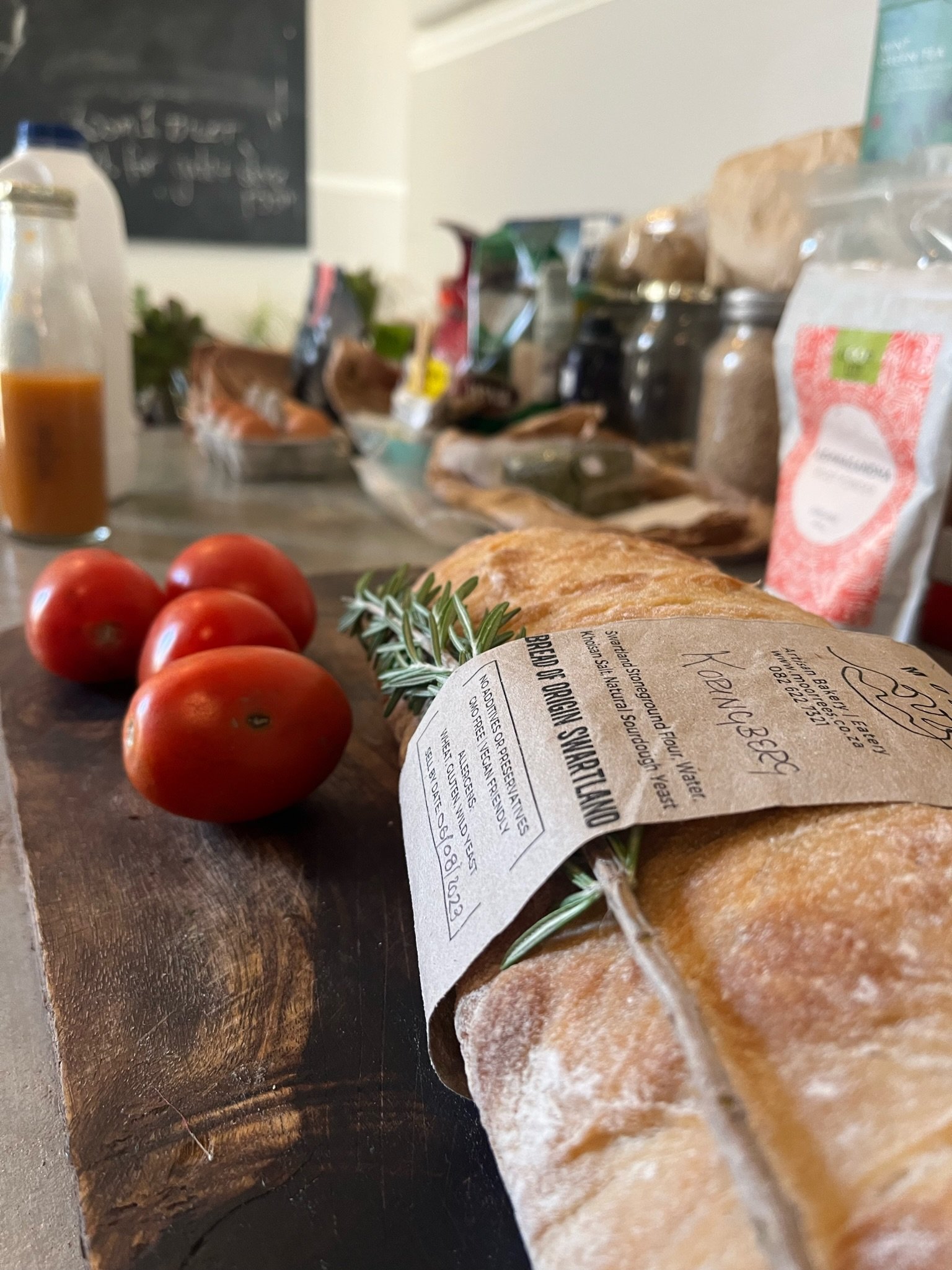 Fresh bread, three ripe tomatoes, and a sprig of rosemary on a rustic wooden surface, with a background of various packaged food items and containers.