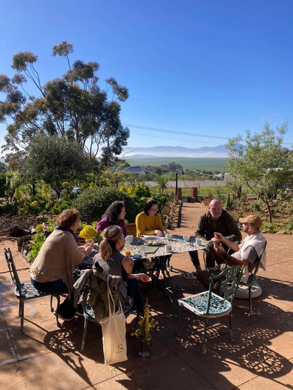 A group of six people sitting around a table outdoors, enjoying a meal on a sunny day with trees and mountains in the background.