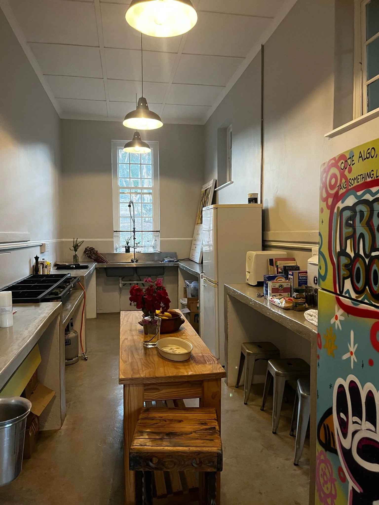 A cozy kitchen with a wooden table and bench, a white refrigerator, a window above the sink, flowers on the table, and various kitchen items on the counter.