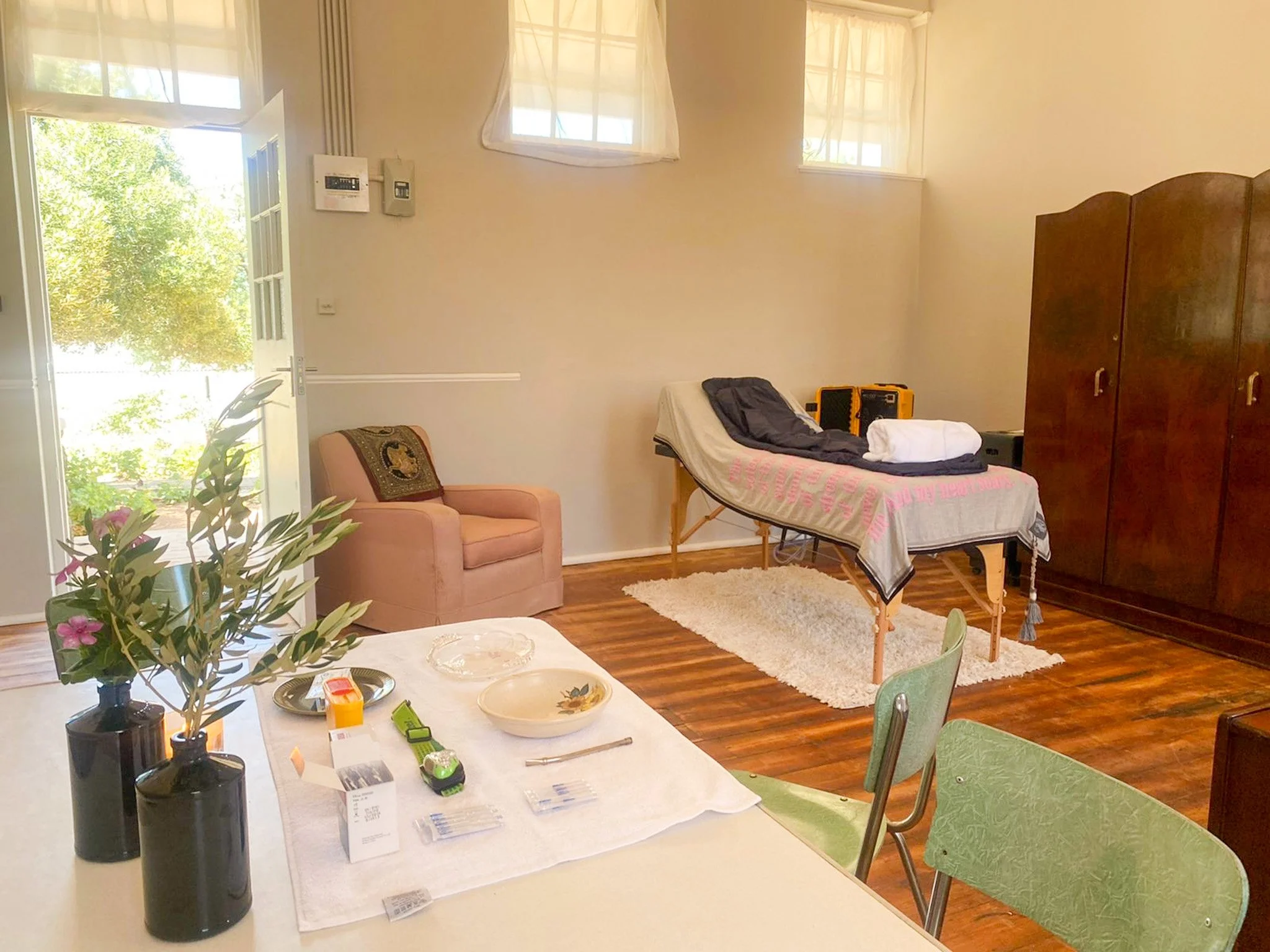 A room with a bed, a pink armchair, a wooden wardrobe, a table with flowers, and an open door leading outside with greenery visible.