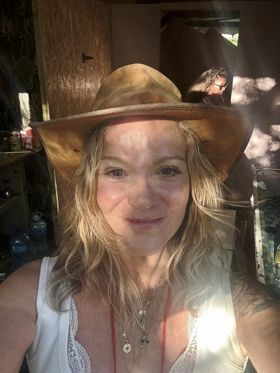 A woman wearing a straw hat and white outfit, sitting outdoors and drinking white wine from a glass, with a bouquet of yellow flowers in front of her.