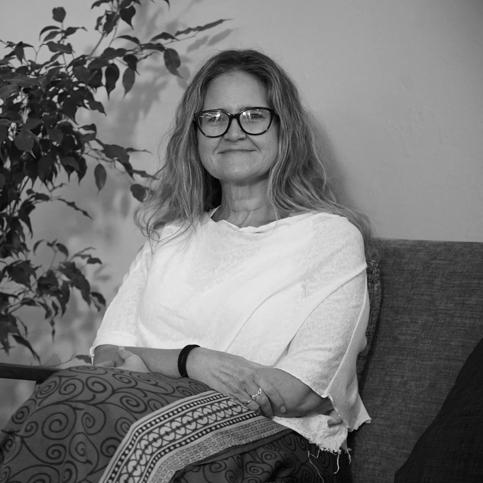 A woman with glasses and wavy hair sitting on a sofa, smiling, with a plant on her left side and a plain wall behind her.