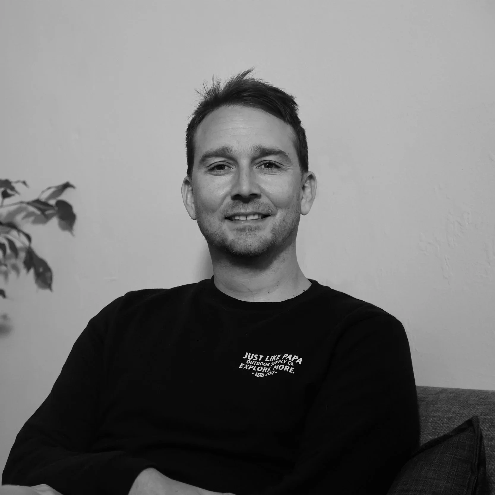 A young man with short hair smiling while sitting on a couch, wearing a black shirt with white text on the chest, in a room with a plain wall and a plant in the background.