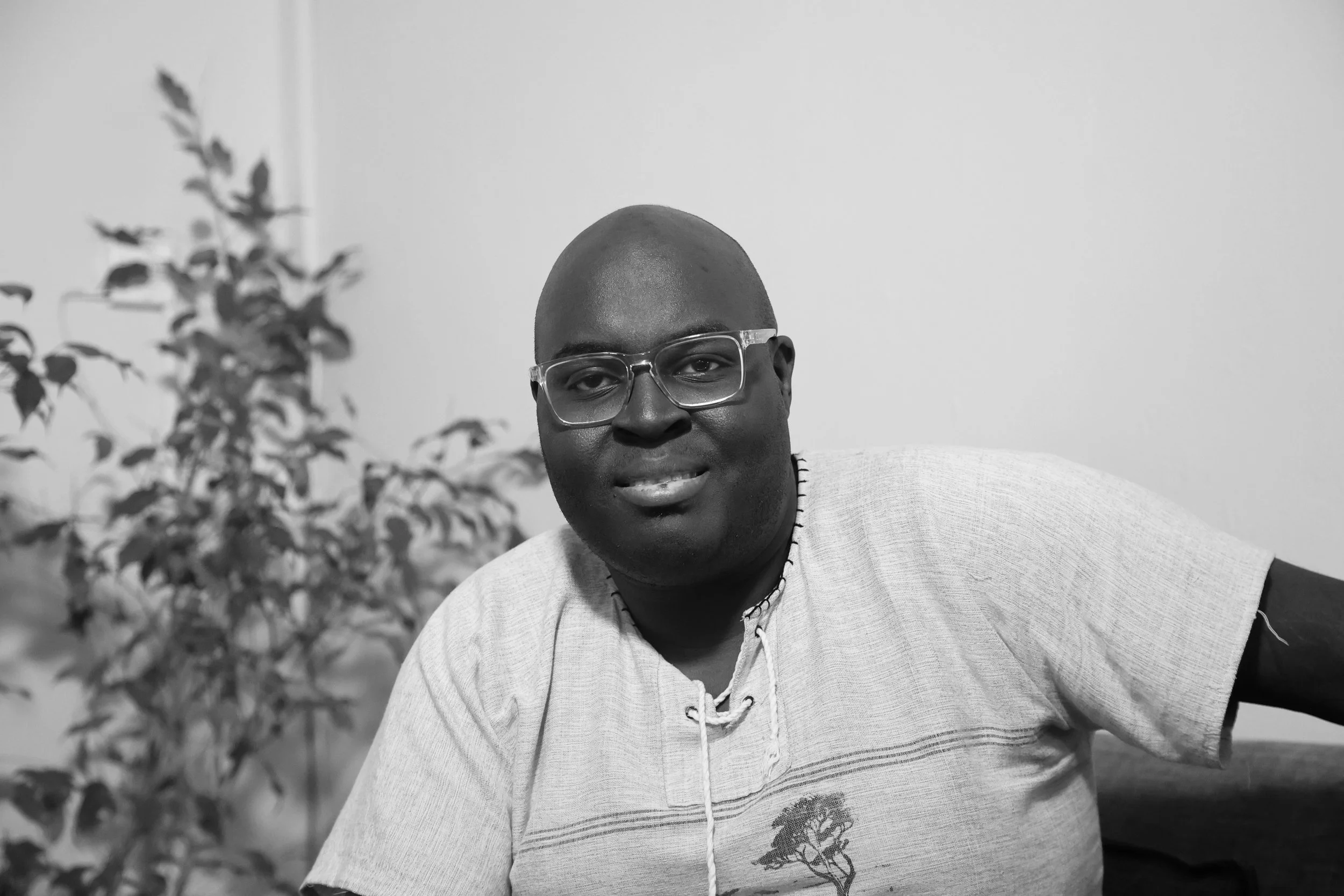 A man with glasses, a shaved head, and a slight smile, wearing a light-colored short-sleeved shirt, sitting indoors with a blurred plant in the background.