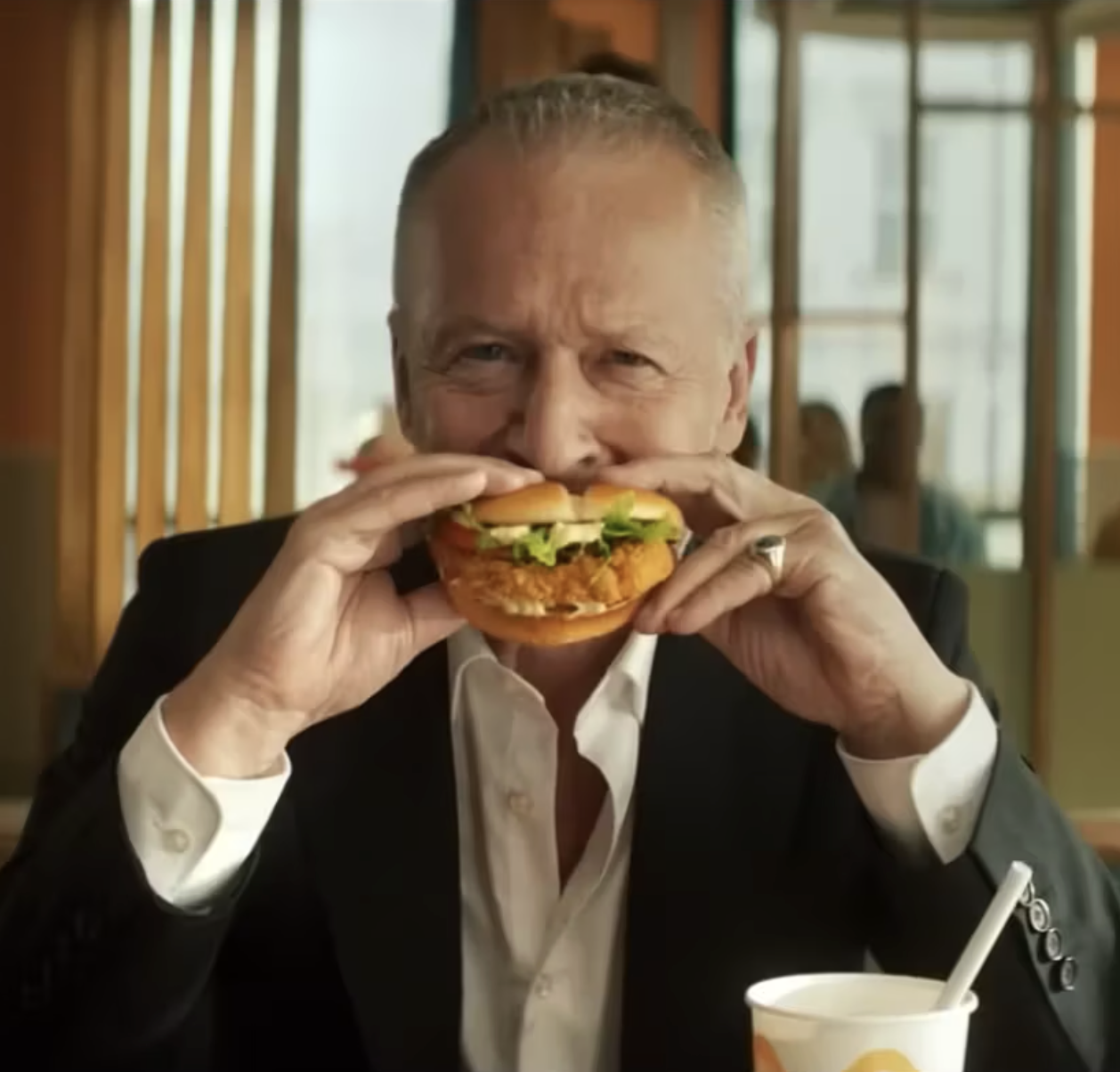 An older man in a dark suit and white shirt is holding a large fried chicken sandwich close to his face and smiling, sitting in a restaurant.