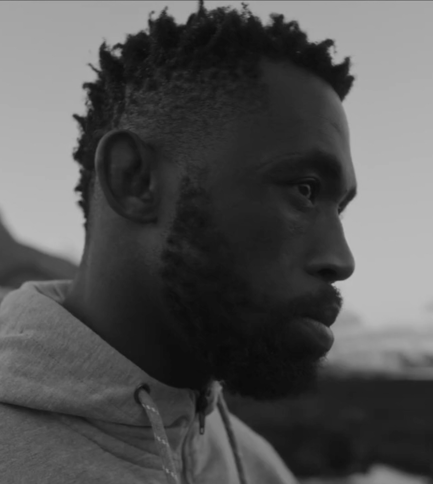 Side profile of a man with a curly haircut, beard, and wearing a hoodie, depicted in black and white.