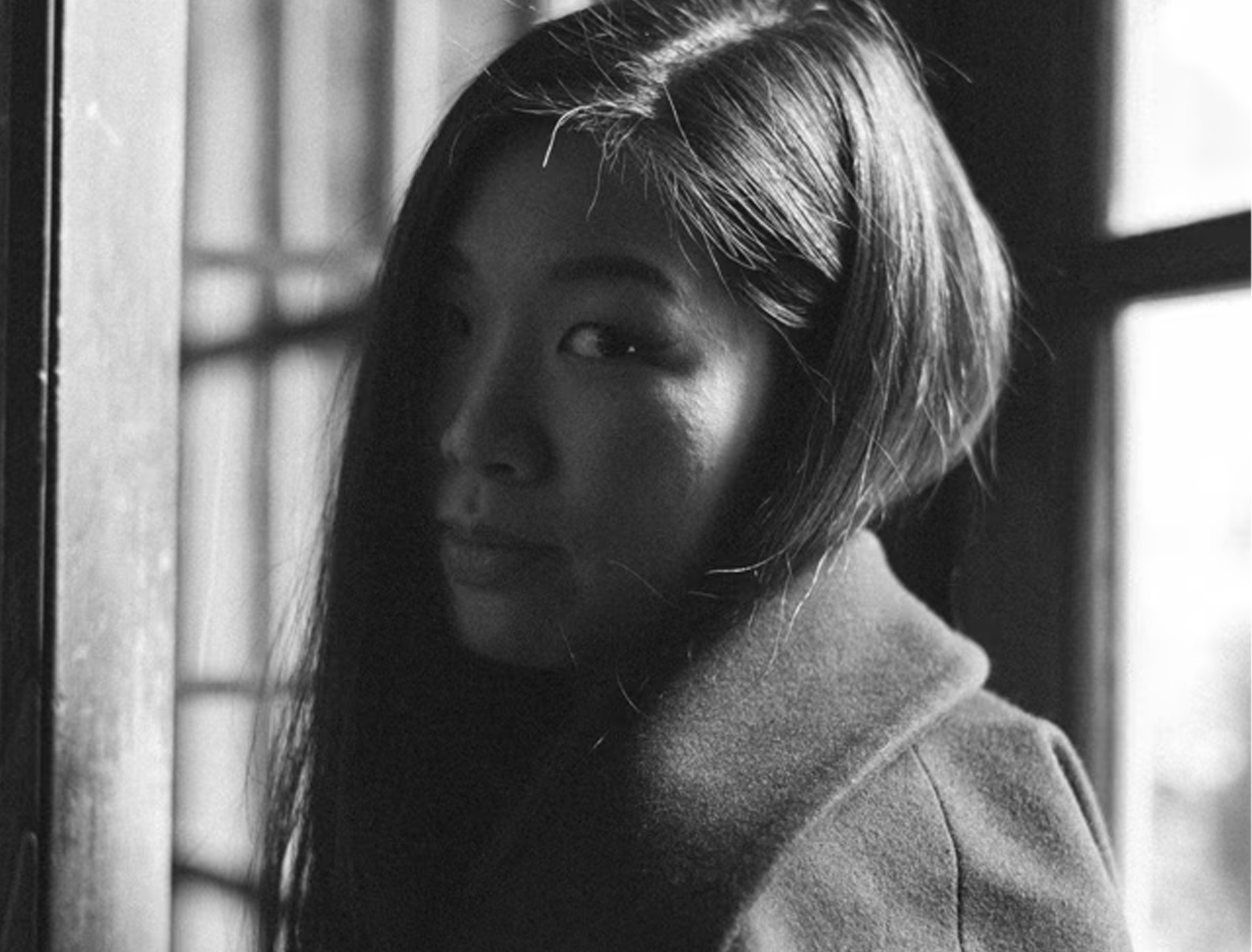 A black-and-white photo of a woman with long dark hair, partly turned to the side, near a window with grid panes, wearing a coat, looking at the camera with a slight smile.