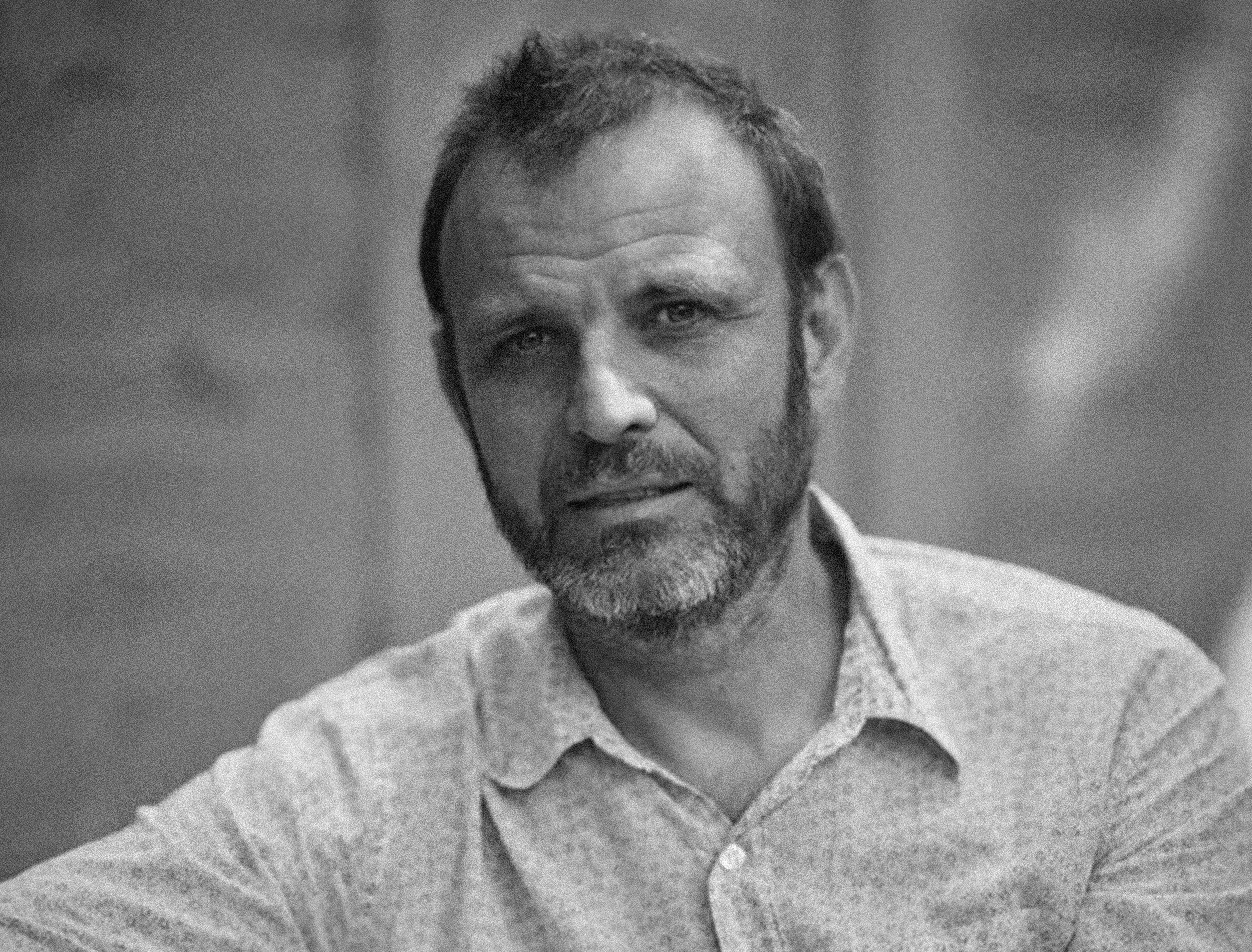 Man with short brown hair, beard, and mustache wearing a light gray button-up shirt, looking directly at the camera with a serious expression.