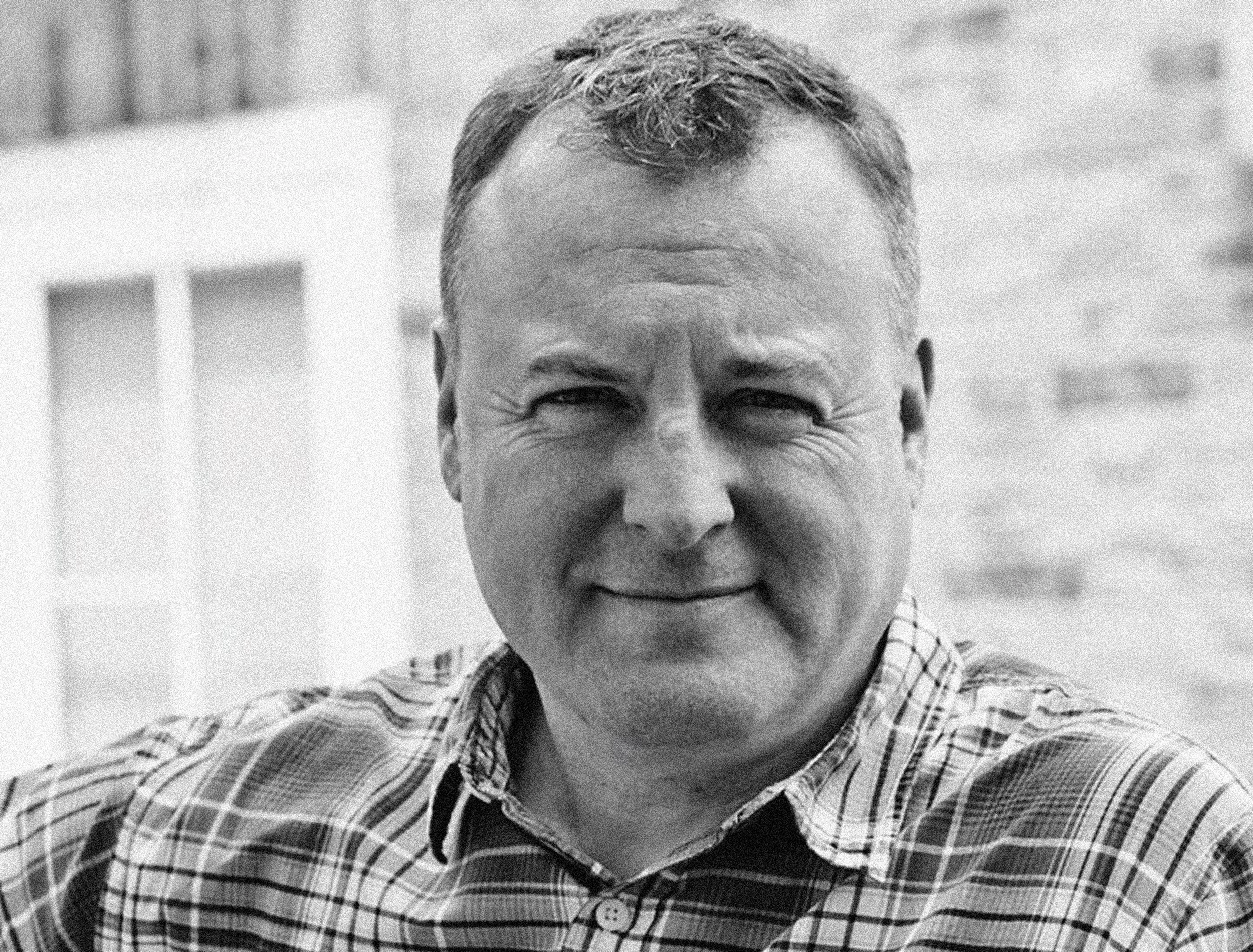 Black and white photo of a middle-aged man smiling, wearing a plaid shirt, outdoors in front of a brick wall and window.