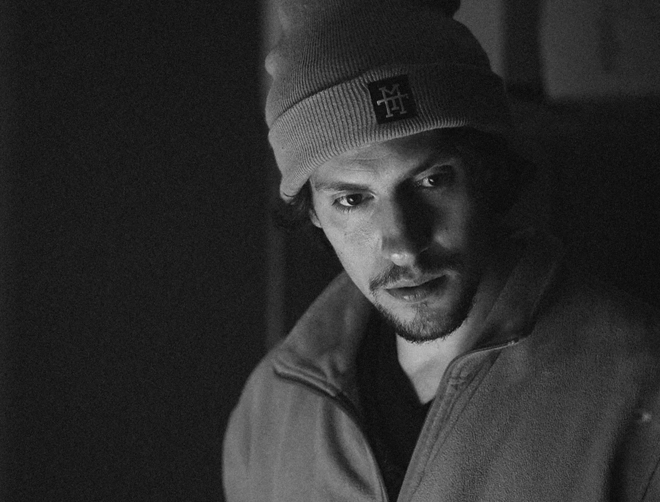 A young man wearing a yellow beanie and an orange jacket, looking at the camera with a serious expression.