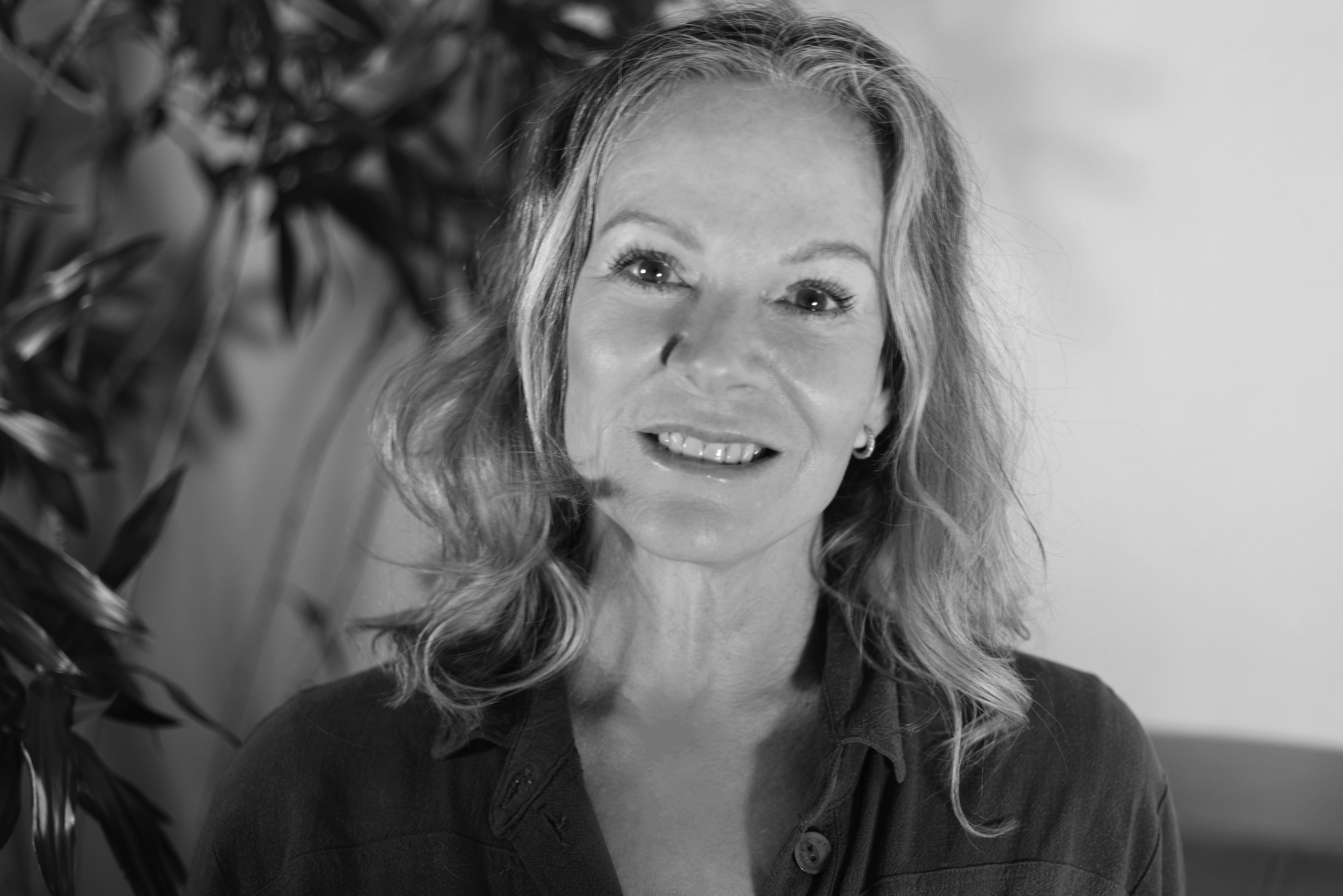 Black and white portrait of a smiling middle-aged woman with shoulder-length wavy hair, wearing a patterned blazer and layered necklaces, standing next to a plant.