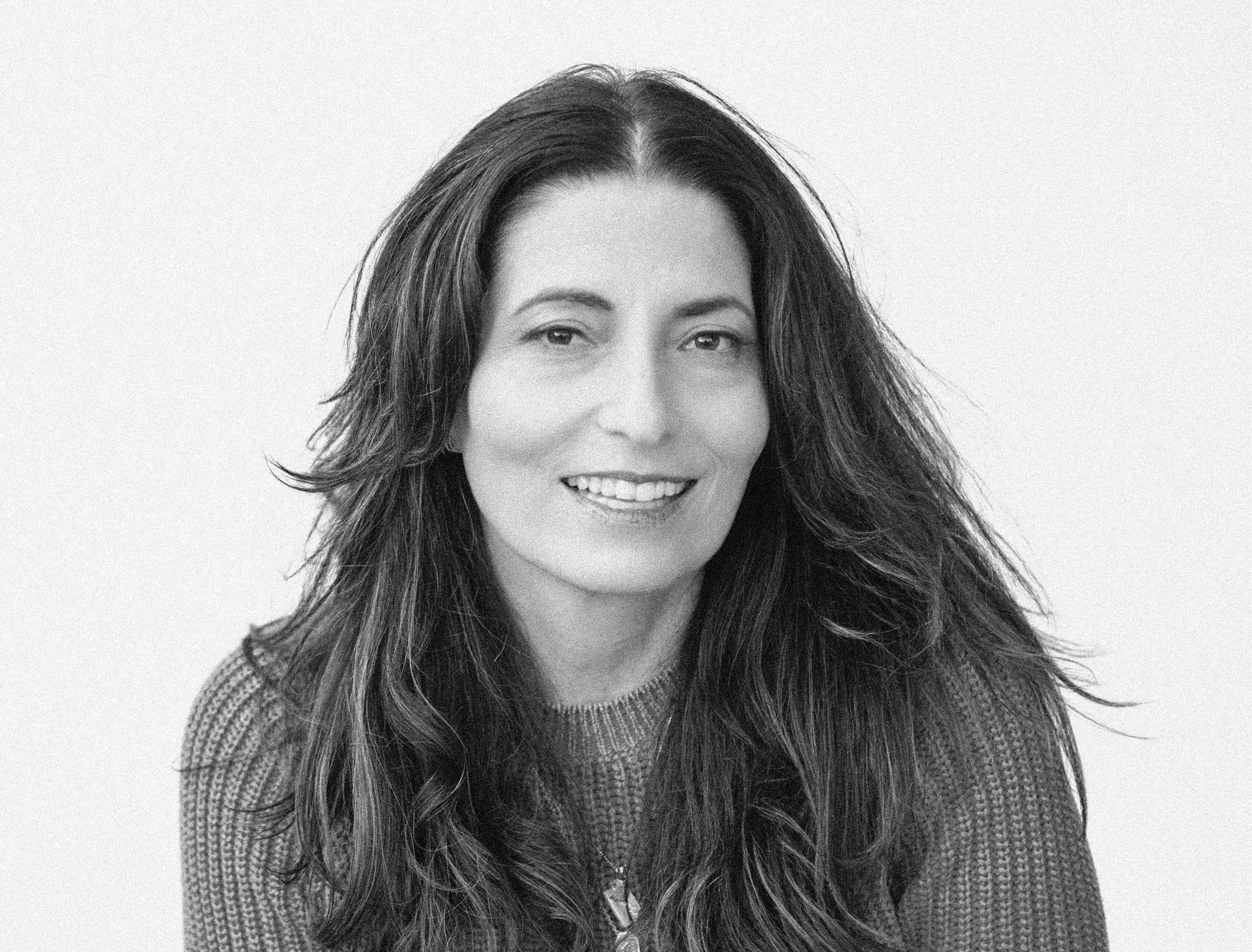 Black and white portrait of a woman with long, wavy hair, smiling with partial teeth showing, wearing a textured sweater and a zipper necklace, against a plain background.