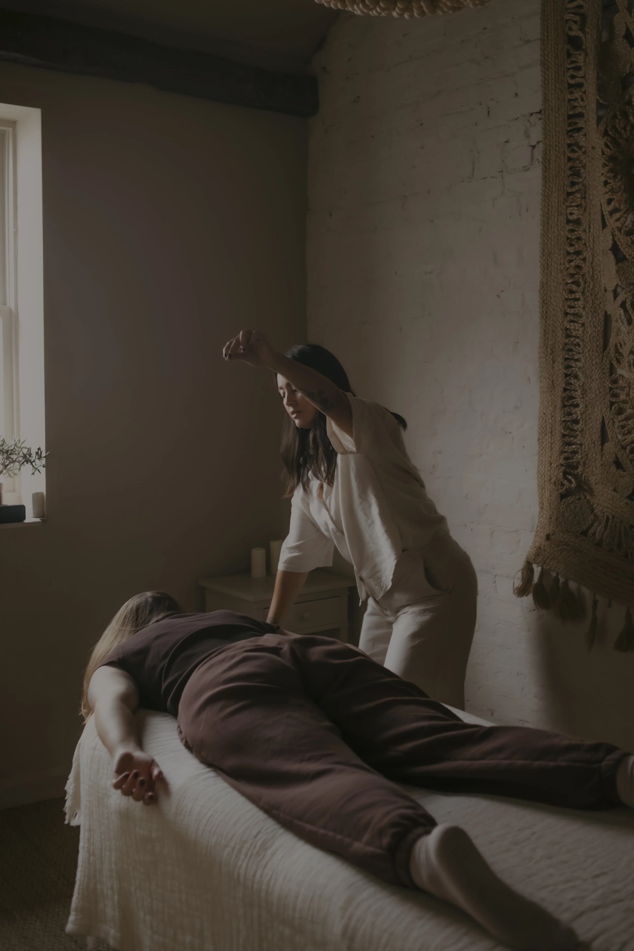 A person is receiving a massage while lying on their stomach on a bed, with a woman standing beside them, reaching over. The room has natural light coming through a window, a white brick wall, and a decorative wall hanging.