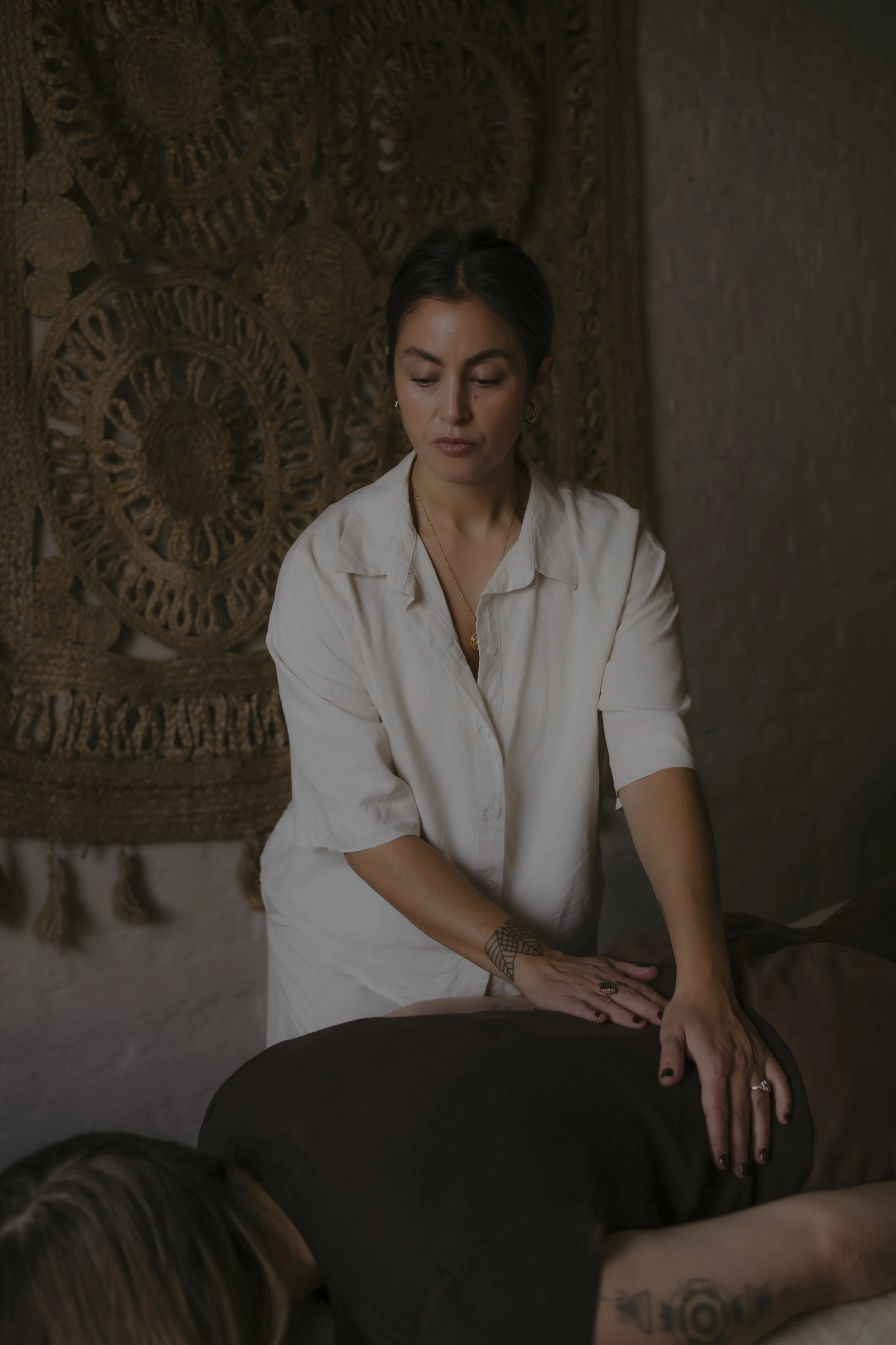 A woman is giving a massage to a person lying face down on a massage table, with a decorative woven wall hanging in the background.