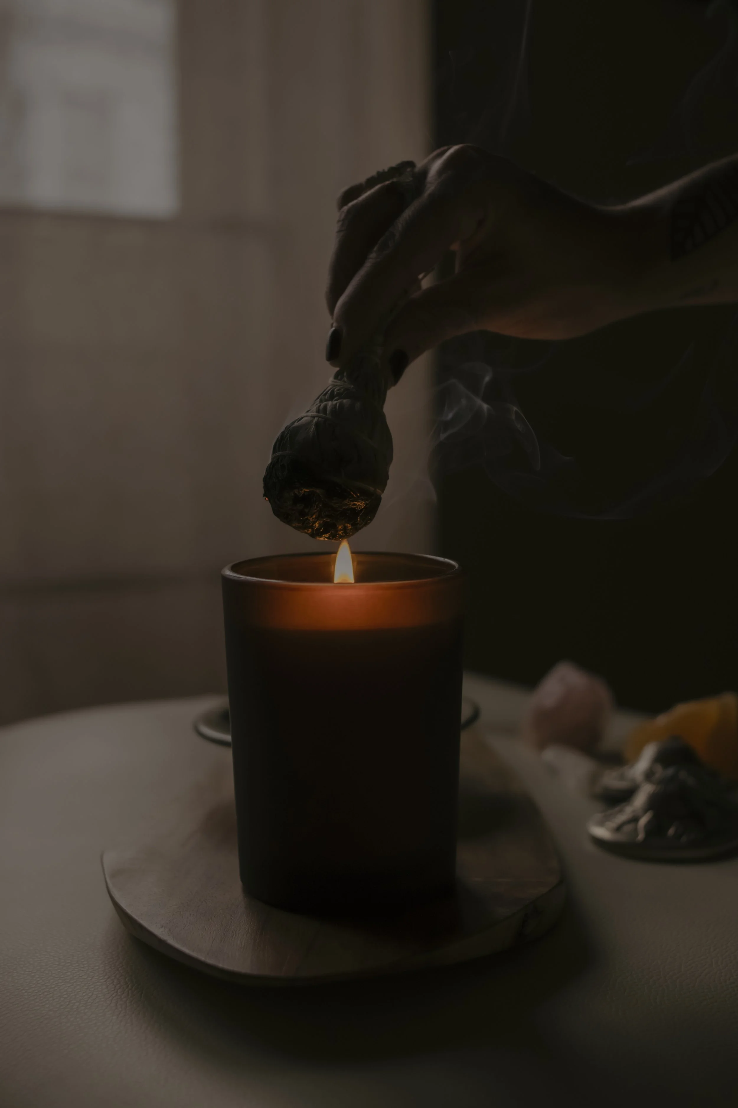 A person holding a large burning incense stick above a lit candle inside a dark room.