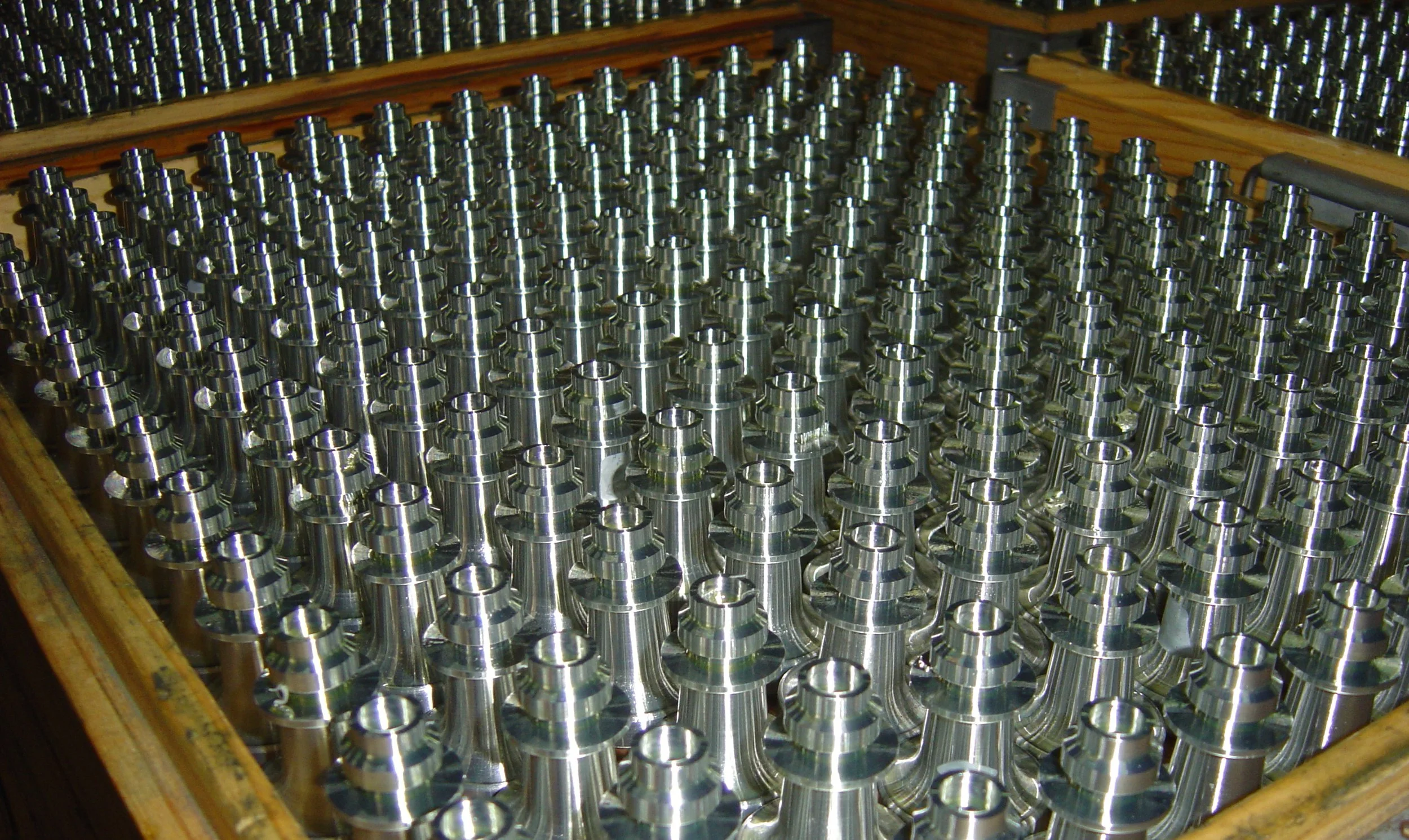 Many shiny, metallic industrial machine parts or components stored in a wooden box.