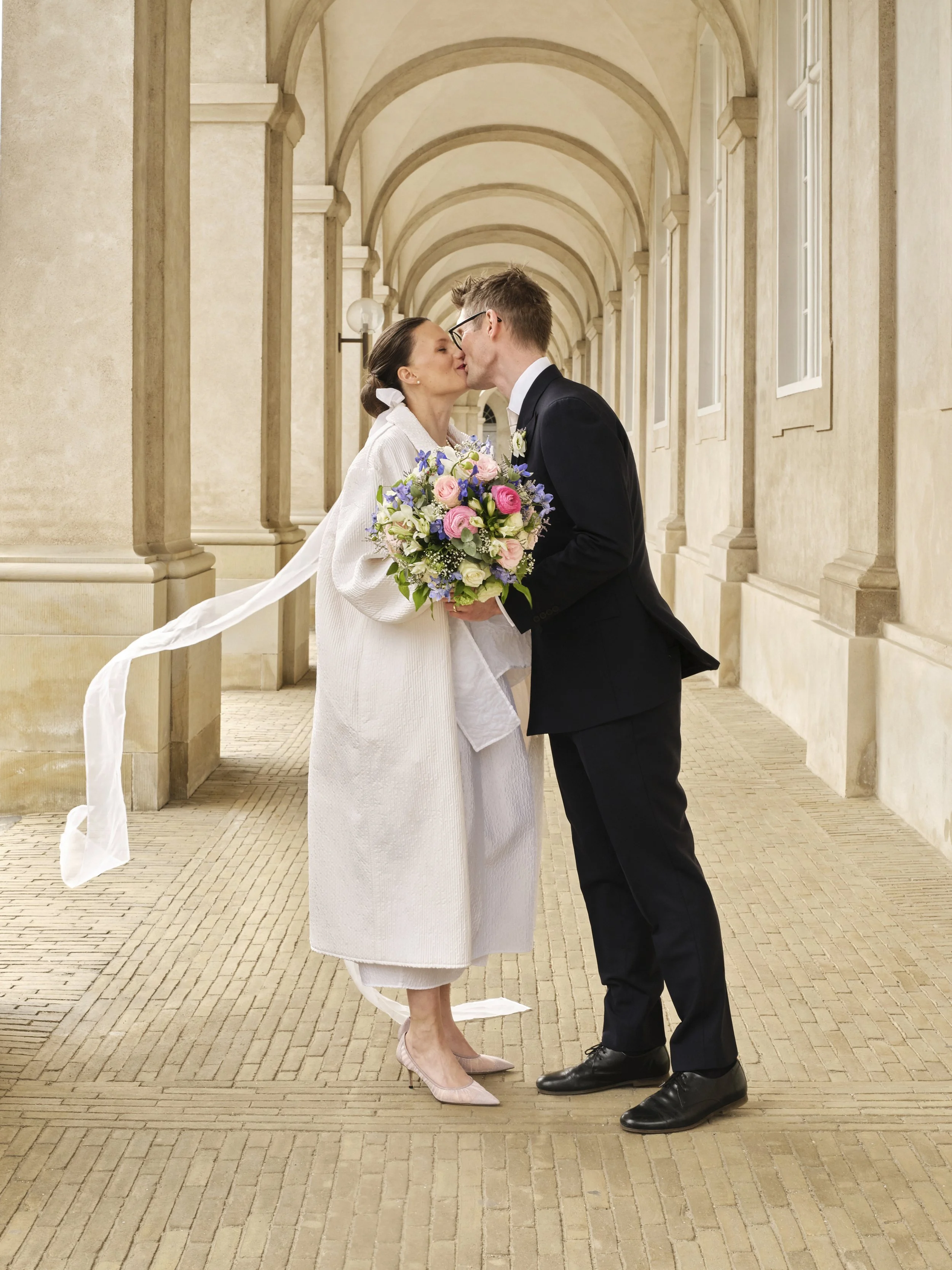 A couple in wedding attire sharing a kiss under an arched hallway. The bride holds a bouquet of pink, purple, and white flowers. The groom wears a black suit and glasses, the bride is dressed in a white coat and heels.