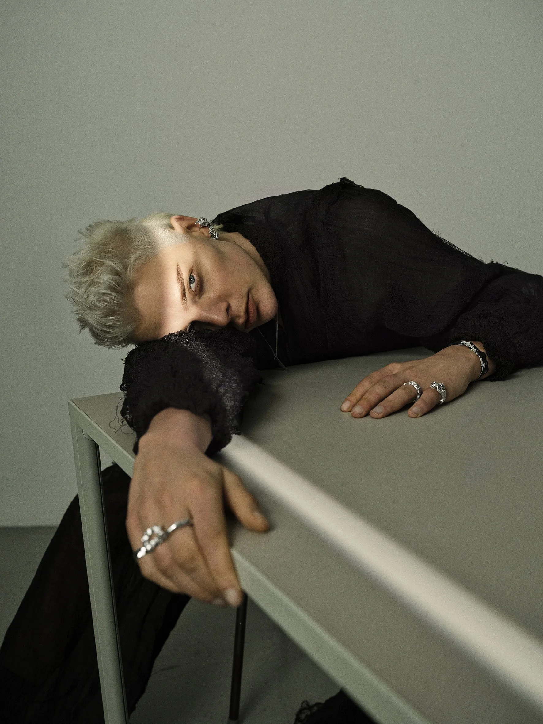 Person with blonde hair lying on a table, resting their head and arm on the surface, wearing black clothing and jewelry. Alice kaufmann jewellery campaign