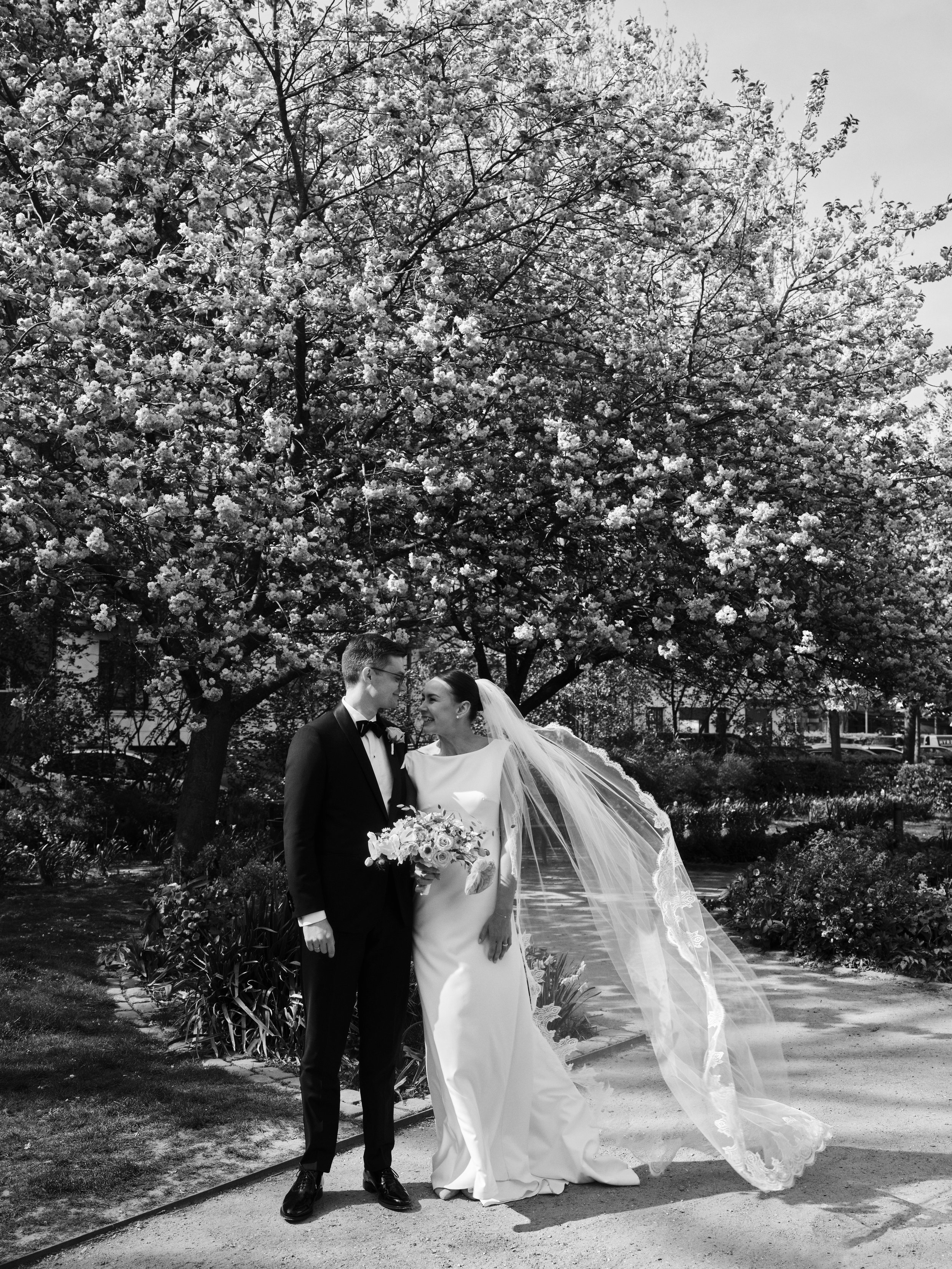 A black and white photo of a bride and groom standing together in a garden, with a large blooming tree in the background. The groom is wearing a tuxedo, and the bride is in a wedding dress with a long veil, holding a bouquet of flowers. They are smiling at each other.