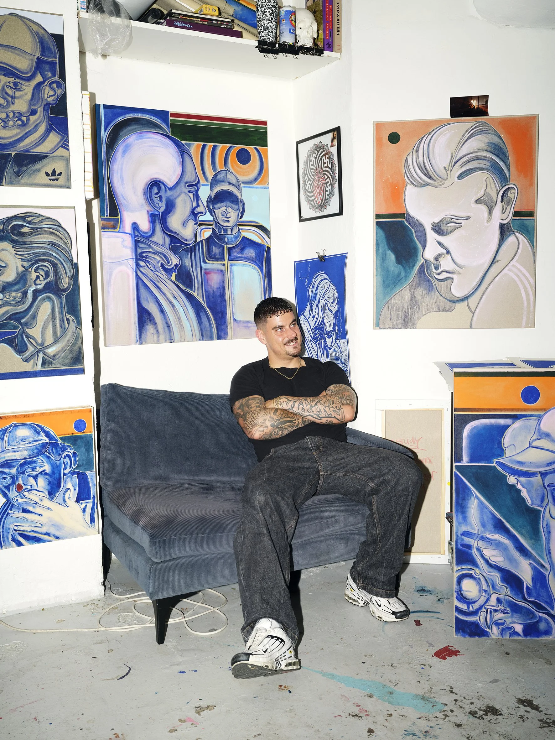 A young man with tattoos on his arms, wearing a black t-shirt and jeans, sits on a gray couch in an art studio. The studio's white walls are decorated with colorful, abstract portrait paintings of various faces, primarily in shades of blue, with some orange and pink accents.