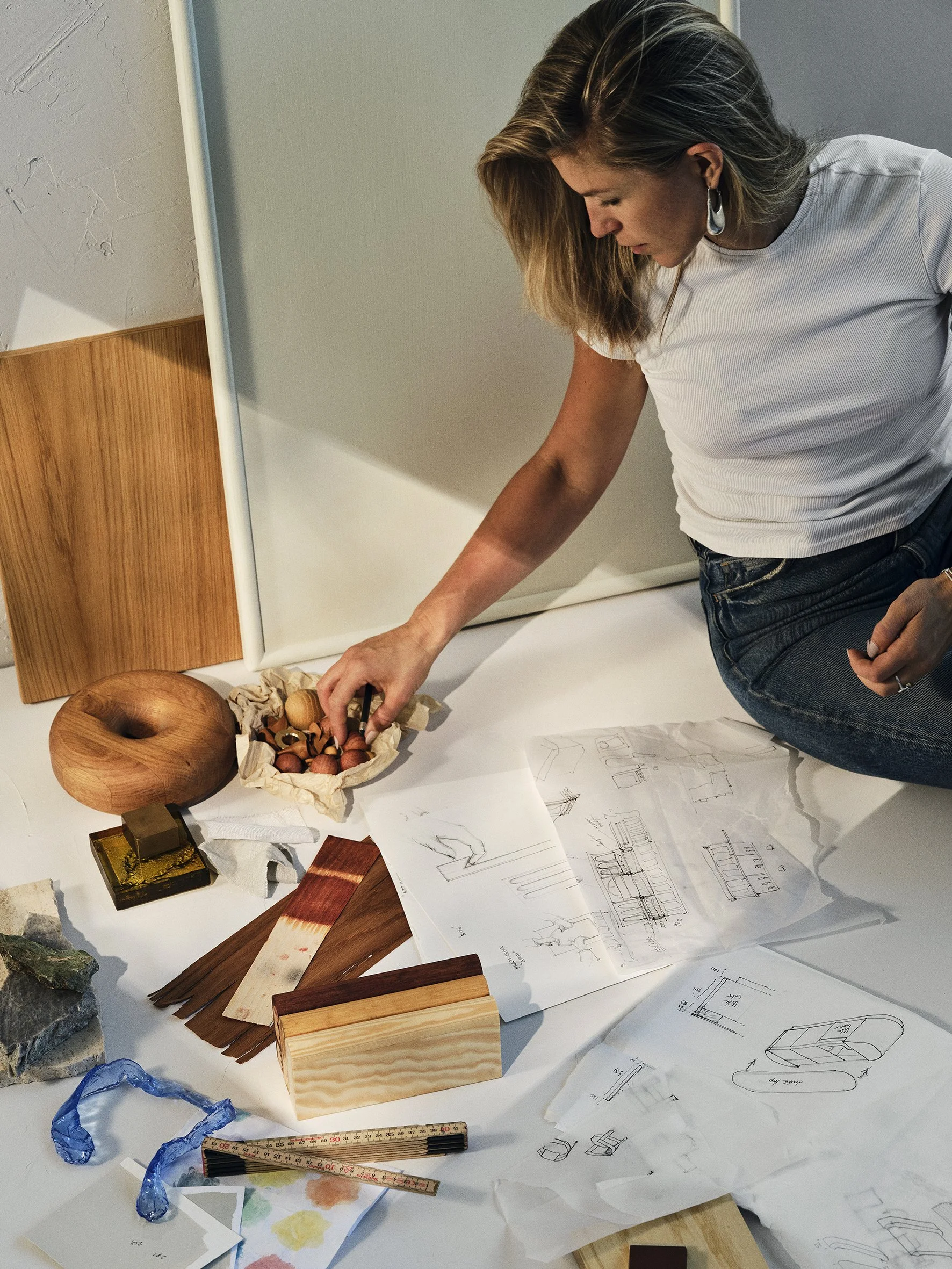 Woman examining wood samples and sketches on a white table for furniture design.