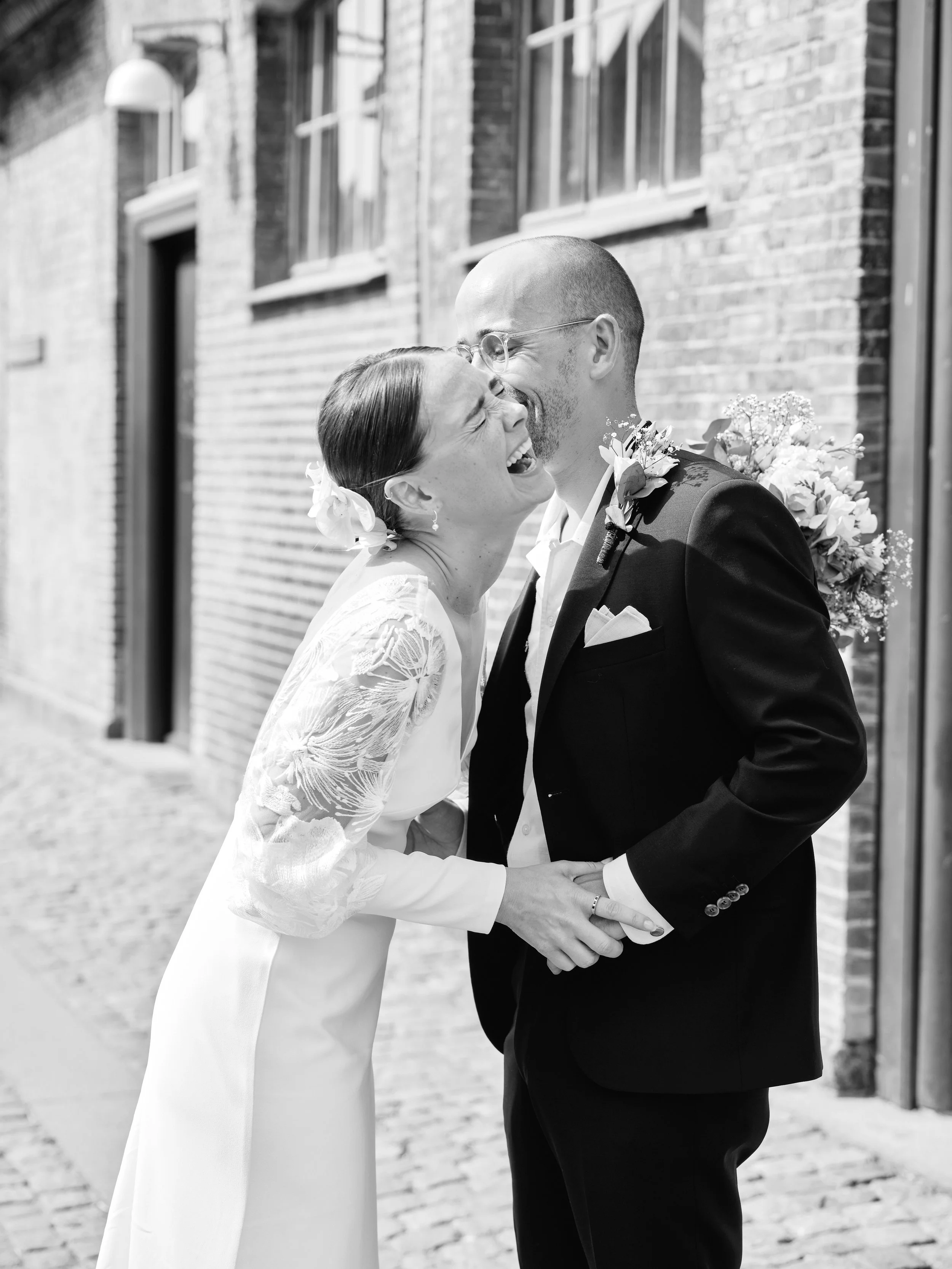 A couple on their wedding day laughing and embracing outdoors against a brick wall, with the bride wearing a lace dress and the groom in a tuxedo.