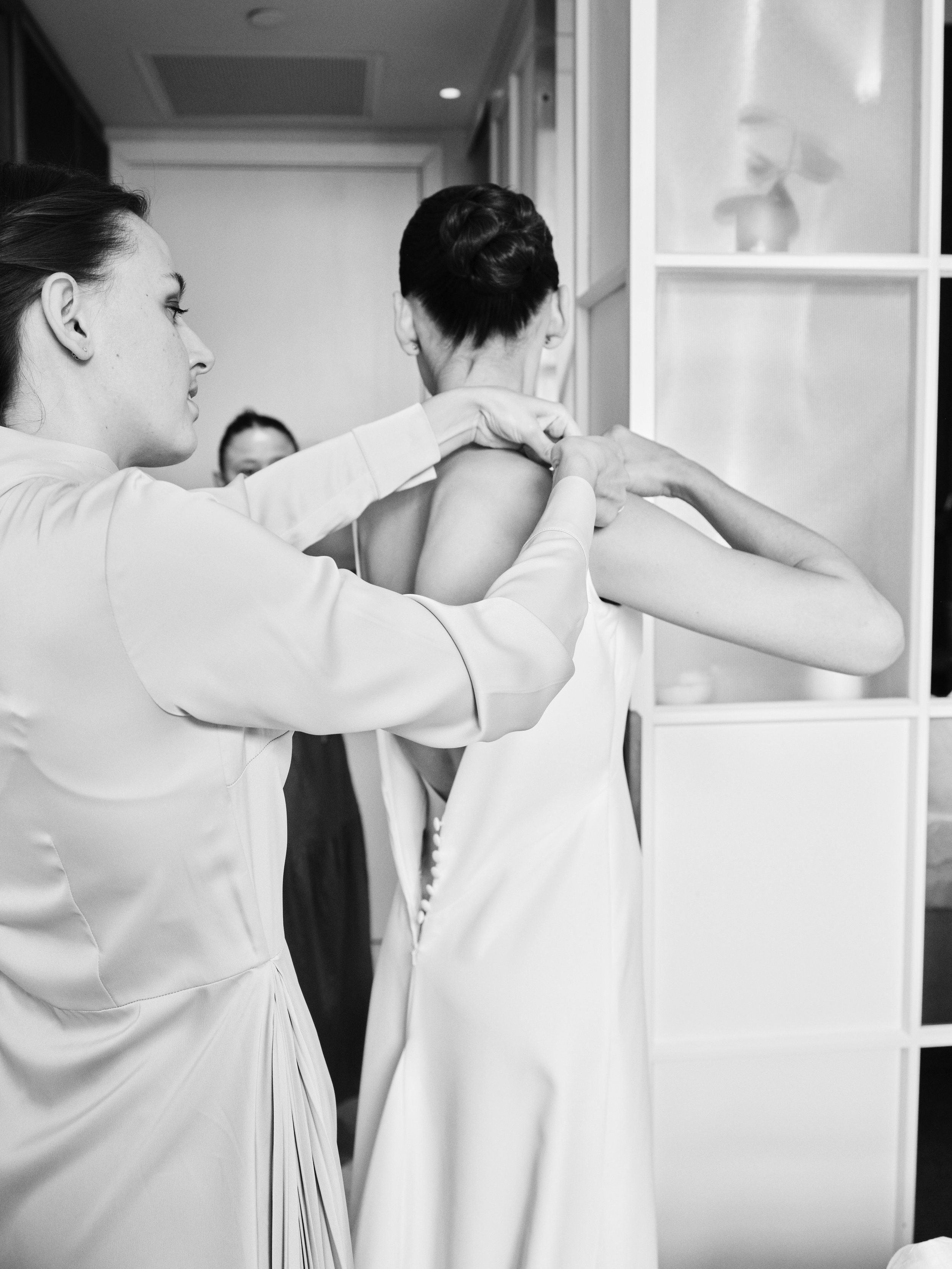 A woman helping another woman put on a dress, likely for a formal event or wedding, in a stylish indoor setting.