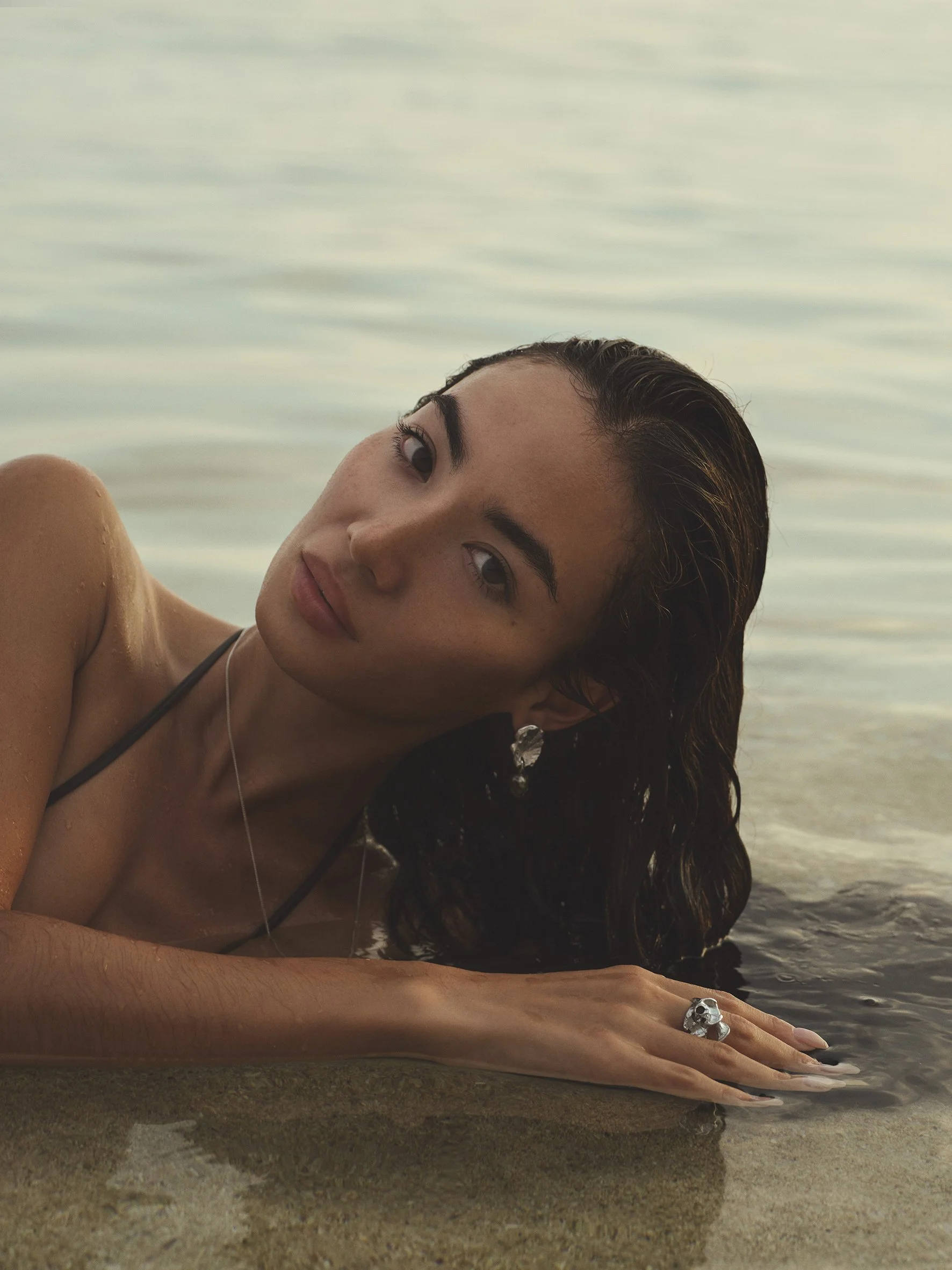 A woman with dark, wet hair lying on a sandy beach near the water, wearing earrings, a ring, and a necklace, looking at the camera.
