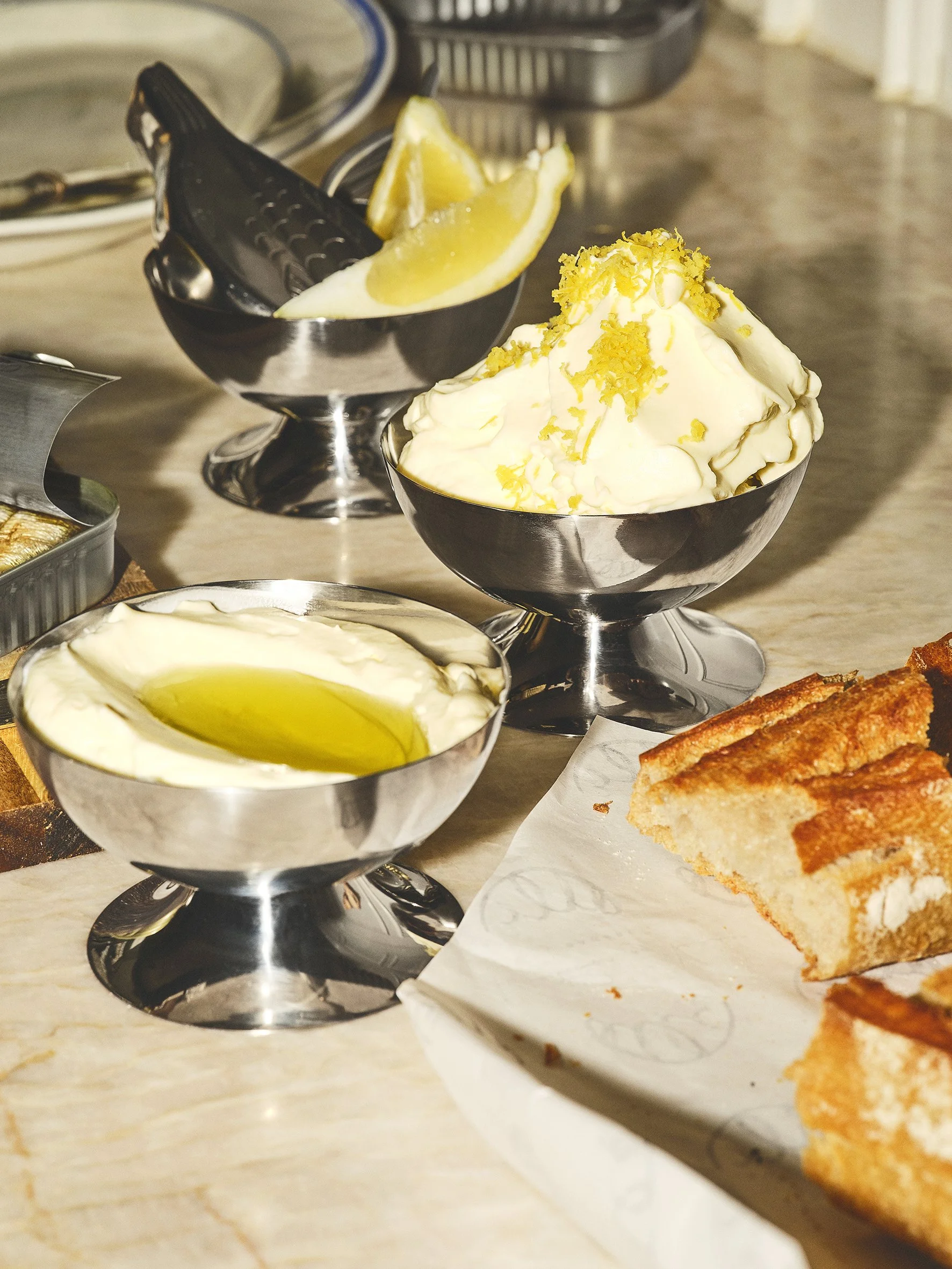 Bowl of lemon wedges and three bowls of lemon curd or pudding on a table, with a slice of cake nearby.