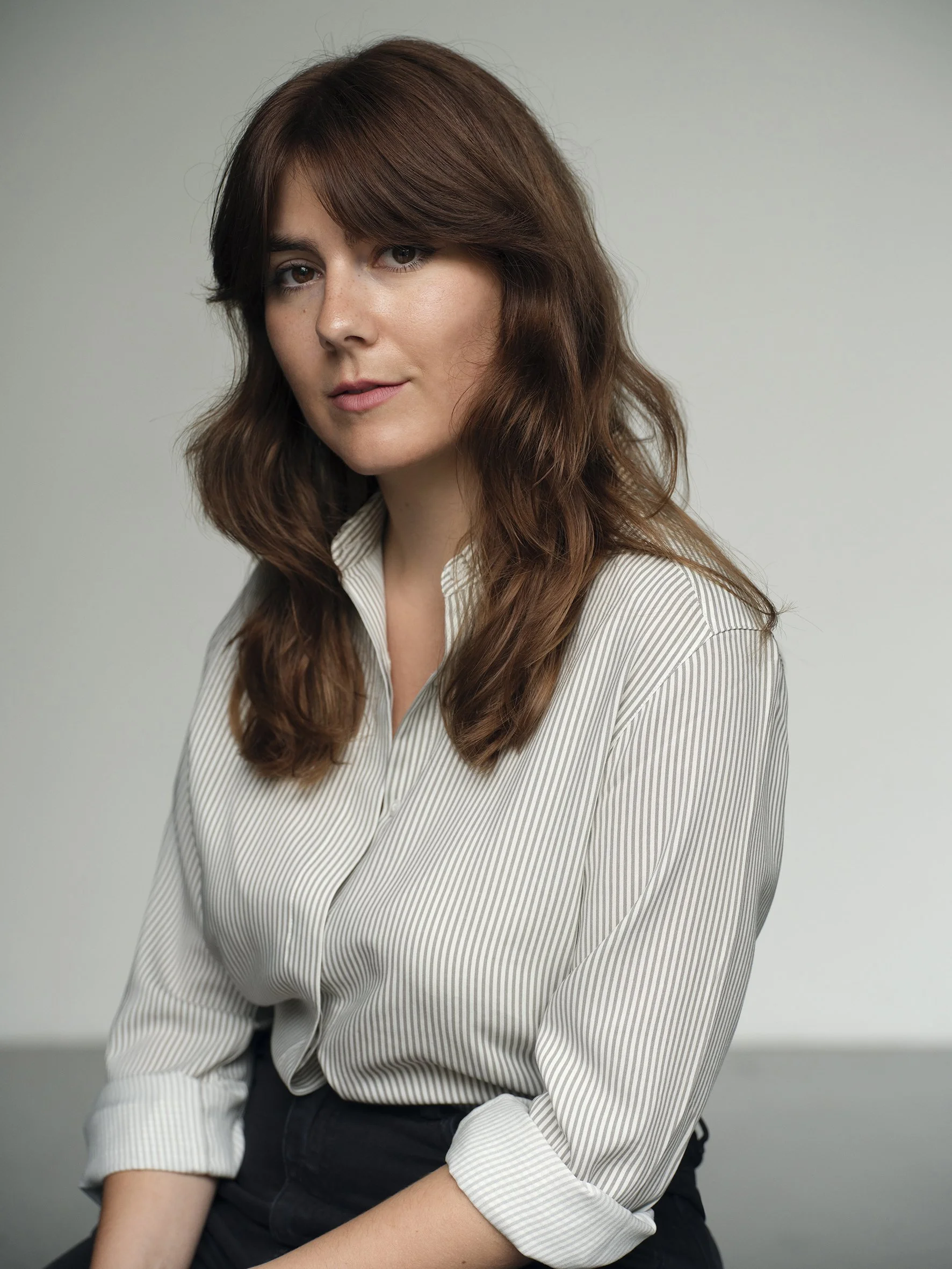 A woman with shoulder-length brown wavy hair, fair skin, wearing a cream-colored striped blouse, sitting in front of a plain light gray background.