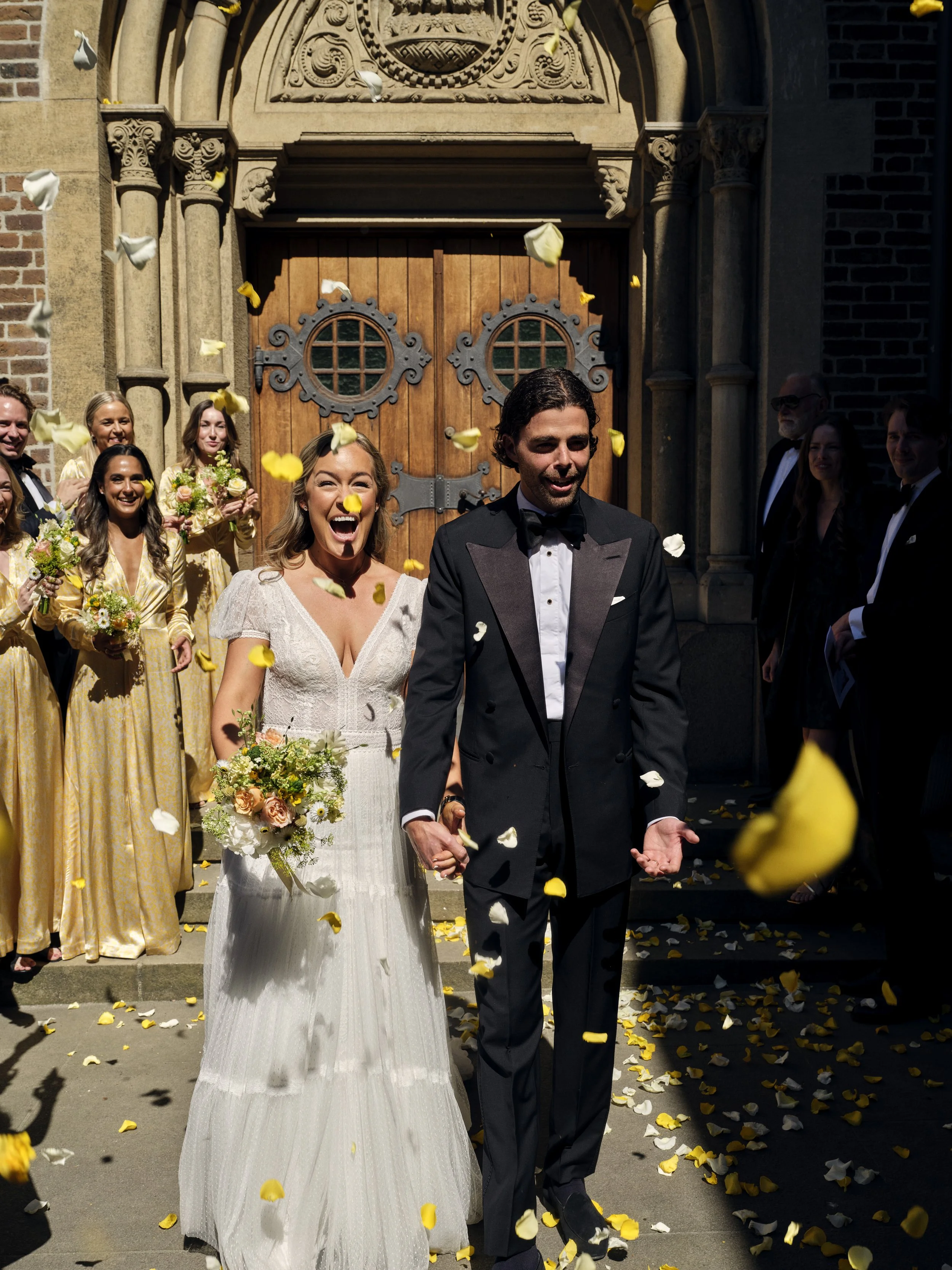 Bride and groom walking out of church holding hands, surrounded by wedding guests throwing flower petals, with church doors in the background.