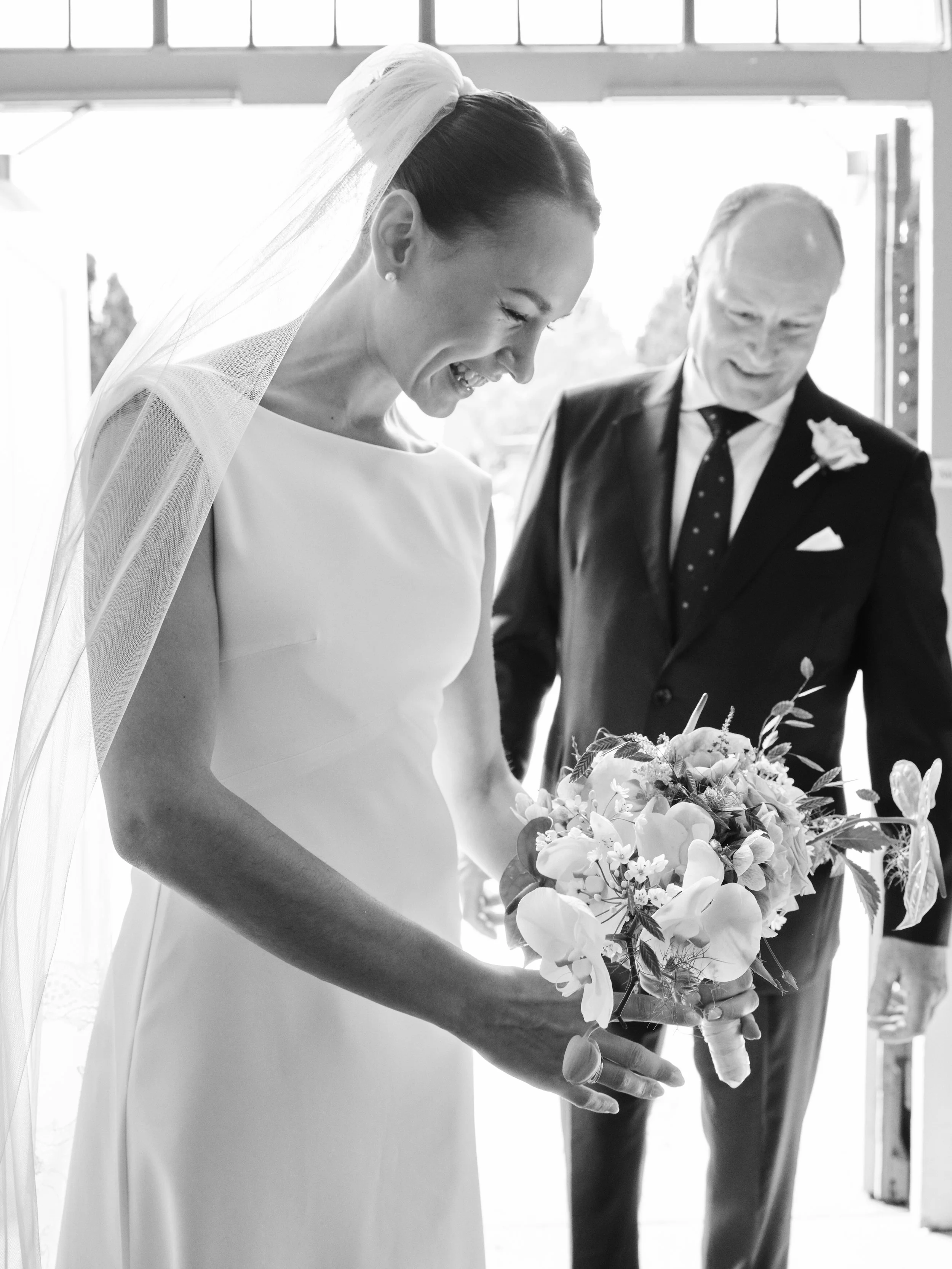 A bride holding a bouquet of flowers, smiling, with an older man in a suit standing behind her, inside a brightly lit room with large windows.