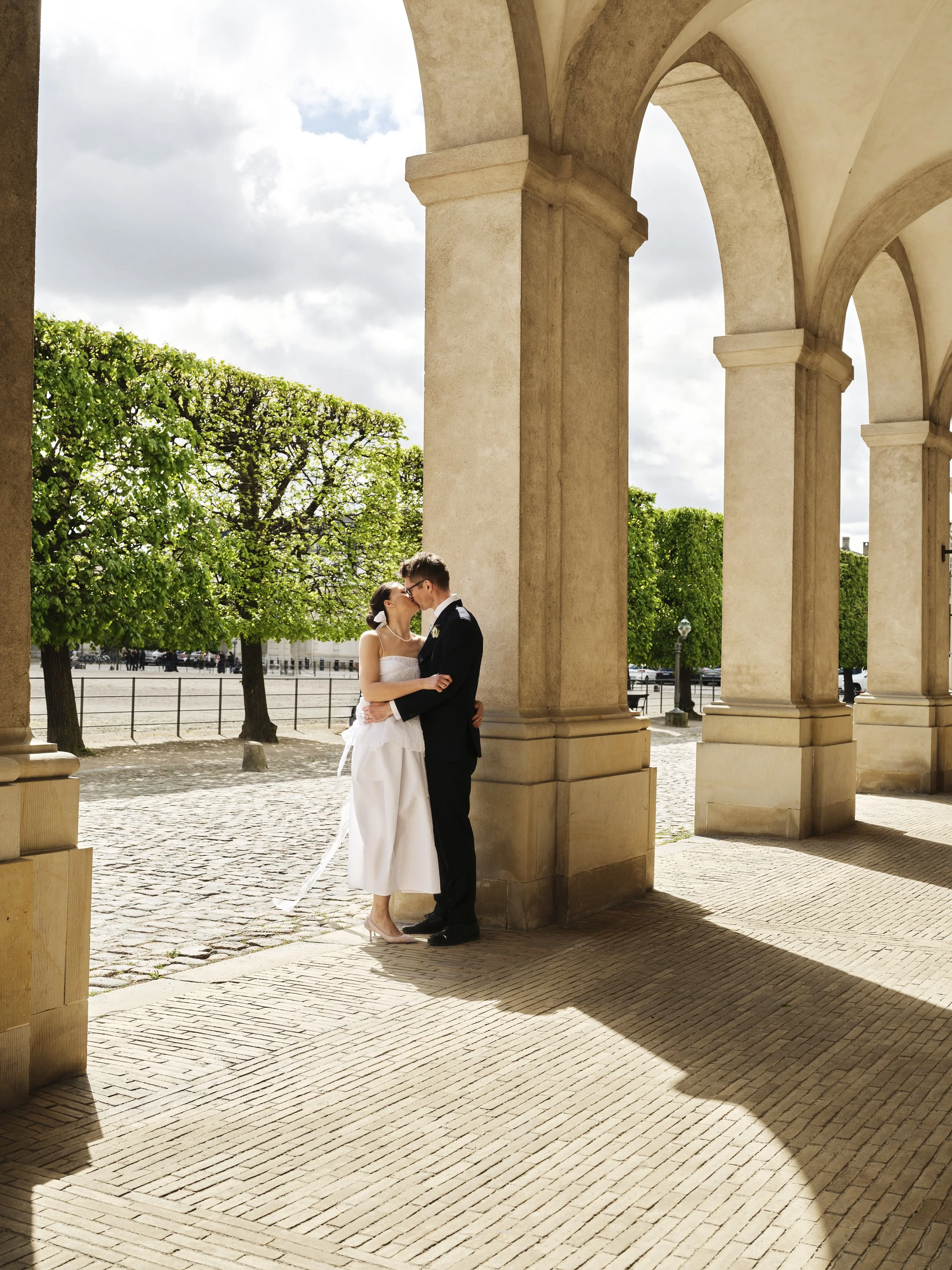 A bride and groom kissing under an archway with stone columns, trees in the background, and cloudy sky.
