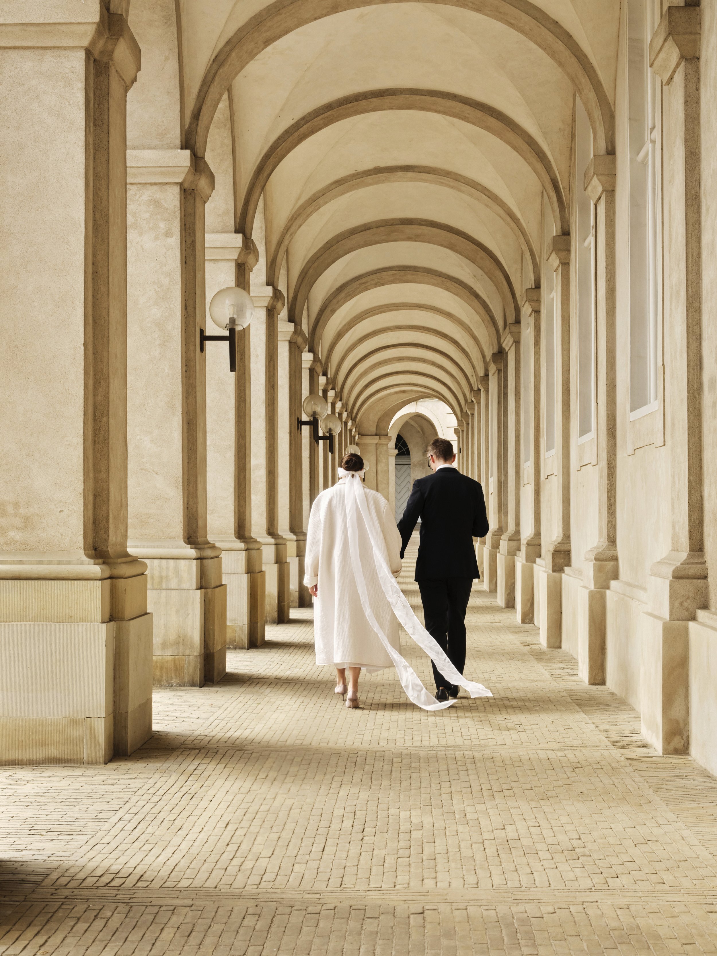 A couple dressed in wedding attire walking hand in hand through an arched colonnade with warm lighting.